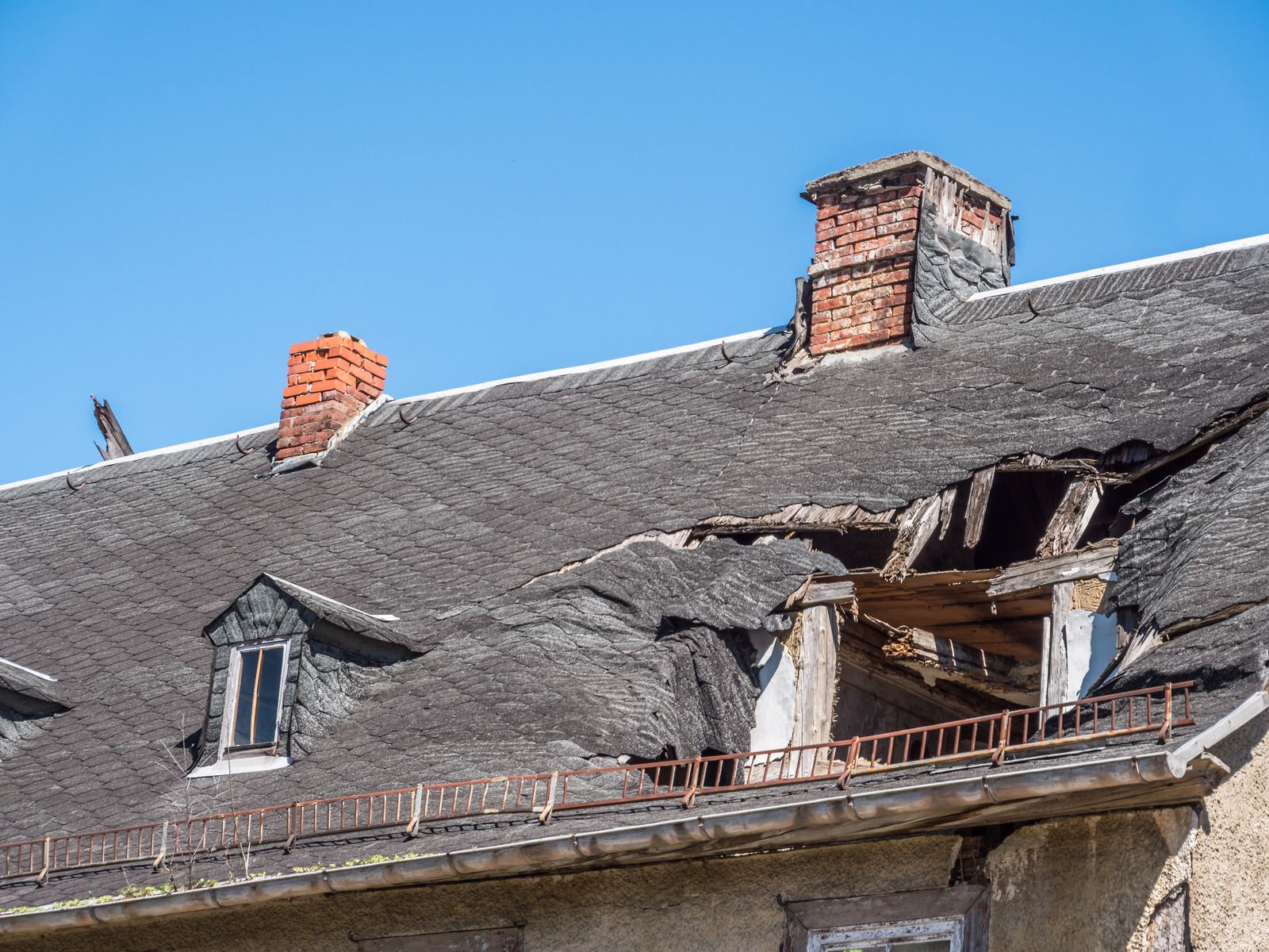 Damaged slate roof on an old building, with a large hole near the chimney against a blue sky