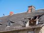 Damaged slate roof on an old building, with a large hole near the chimney against a blue sky