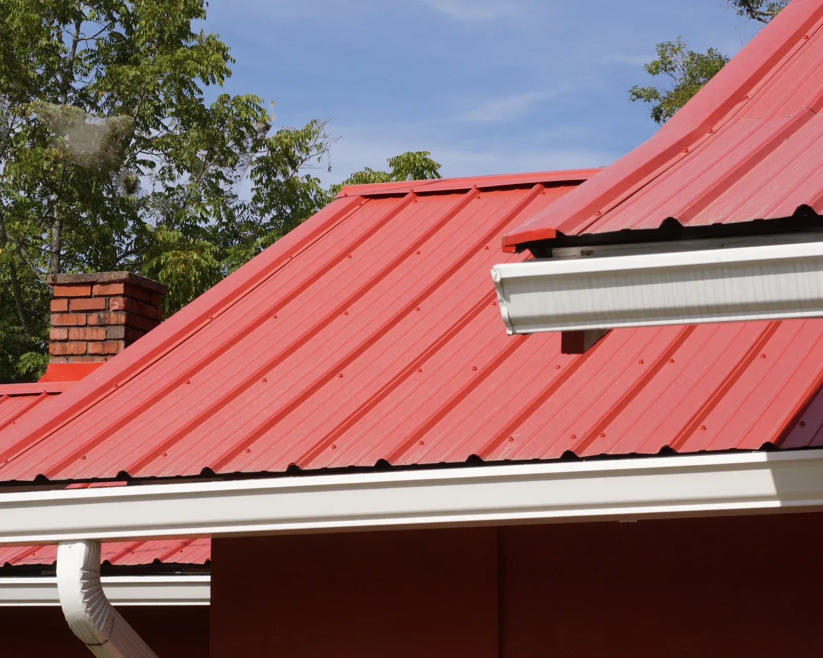 Red metal roof with white gutters and a brick chimney against trees and blue sky