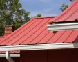 Red metal roof with white gutters and a brick chimney against trees and blue sky