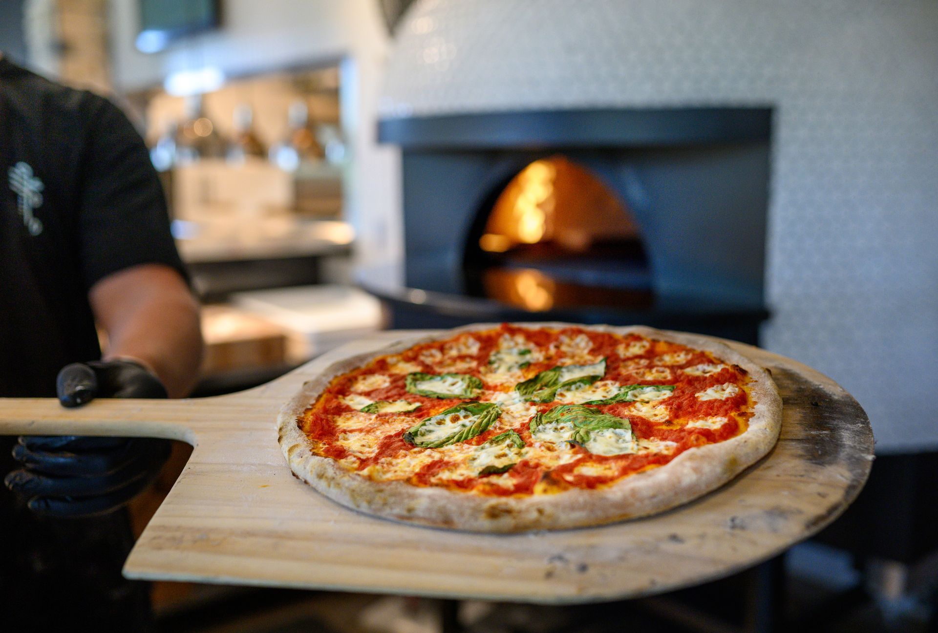 A person is holding a pizza on a wooden paddle in front of a pizza oven.