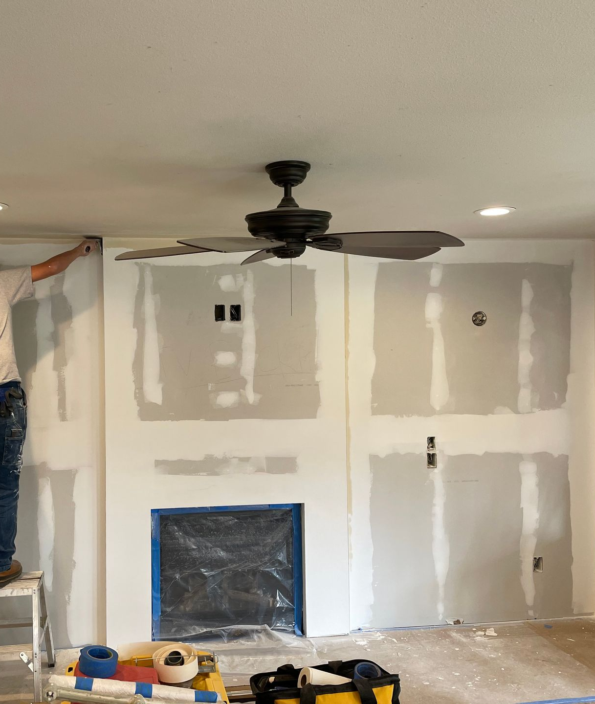 Living room under renovation with unfinished drywall, a ceiling fan, and tools on the floor.