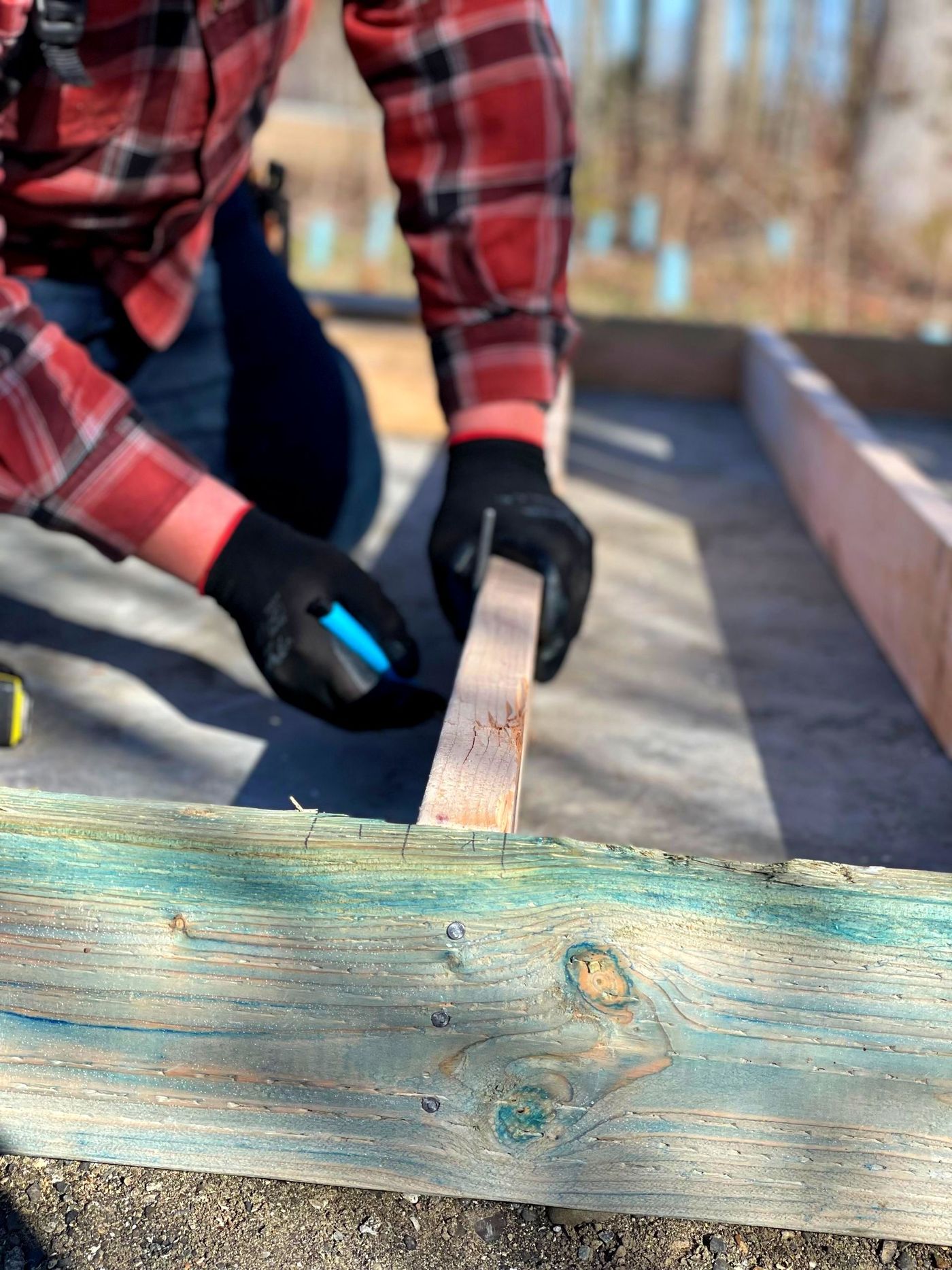 Person in red plaid shirt measuring a wooden plank on an outdoor construction project