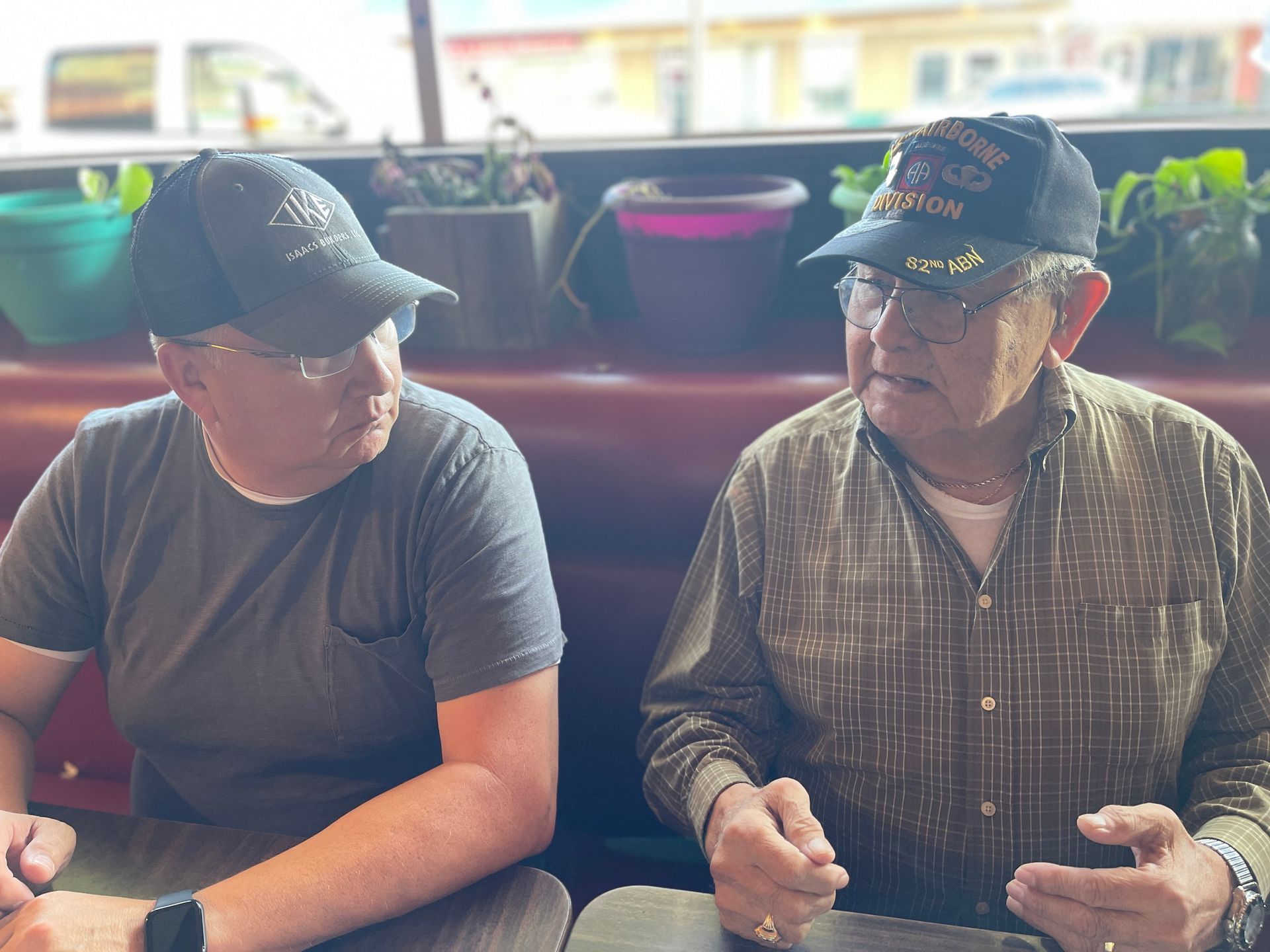 Two people in baseball caps sit at a café table, chatting near a window with potted plants.