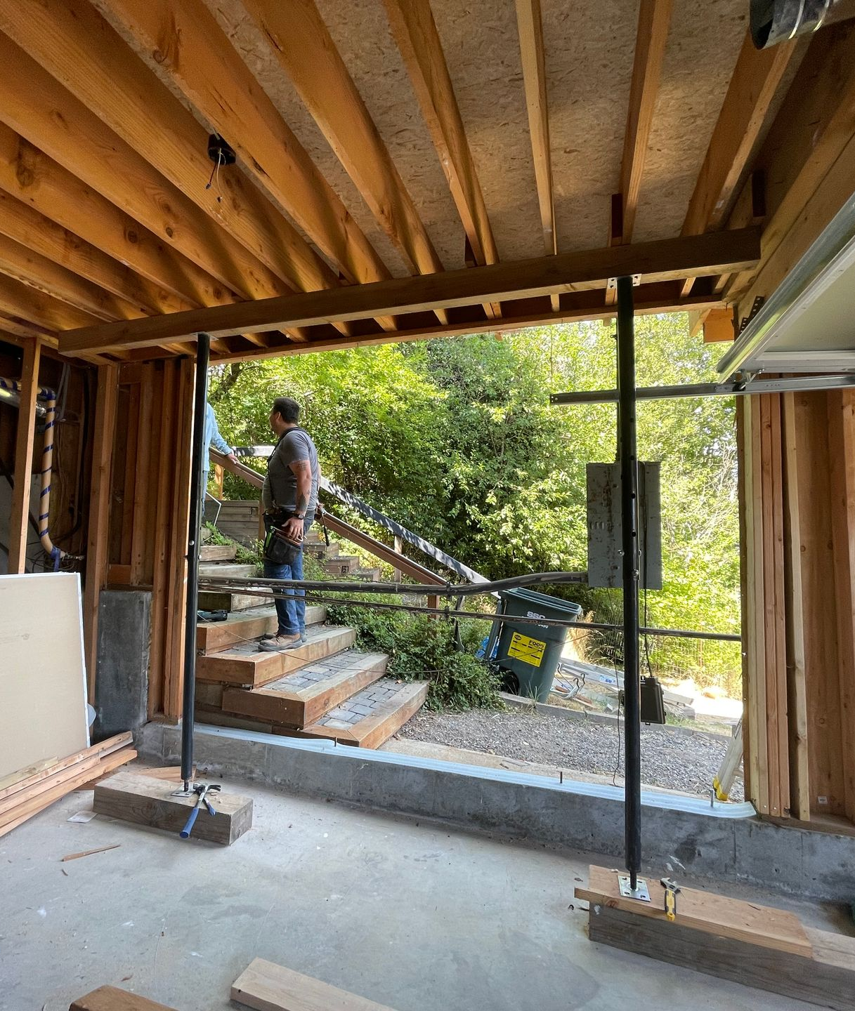 Unfinished room opening to stairs and greenery outside, with a worker standing near the steps.