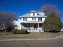 White two-story house with porch and dormer, under a bright blue sky. Trees surround.
