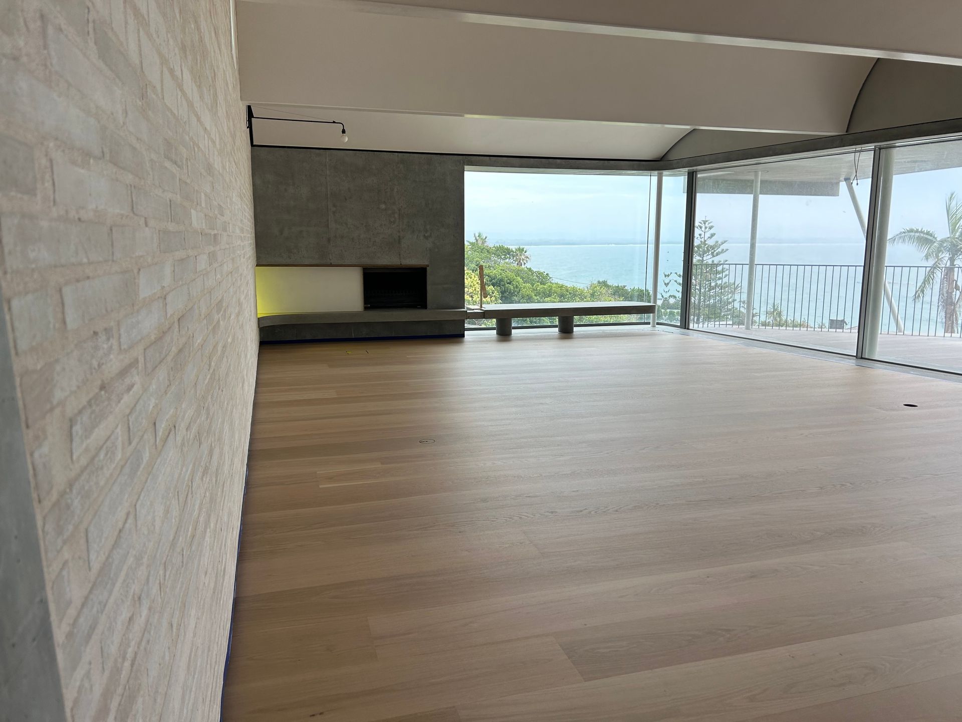 Empty room with light wood floors, a brick wall, and a view of the ocean through large windows — CM Timber Flooring in East Ballina, NSW