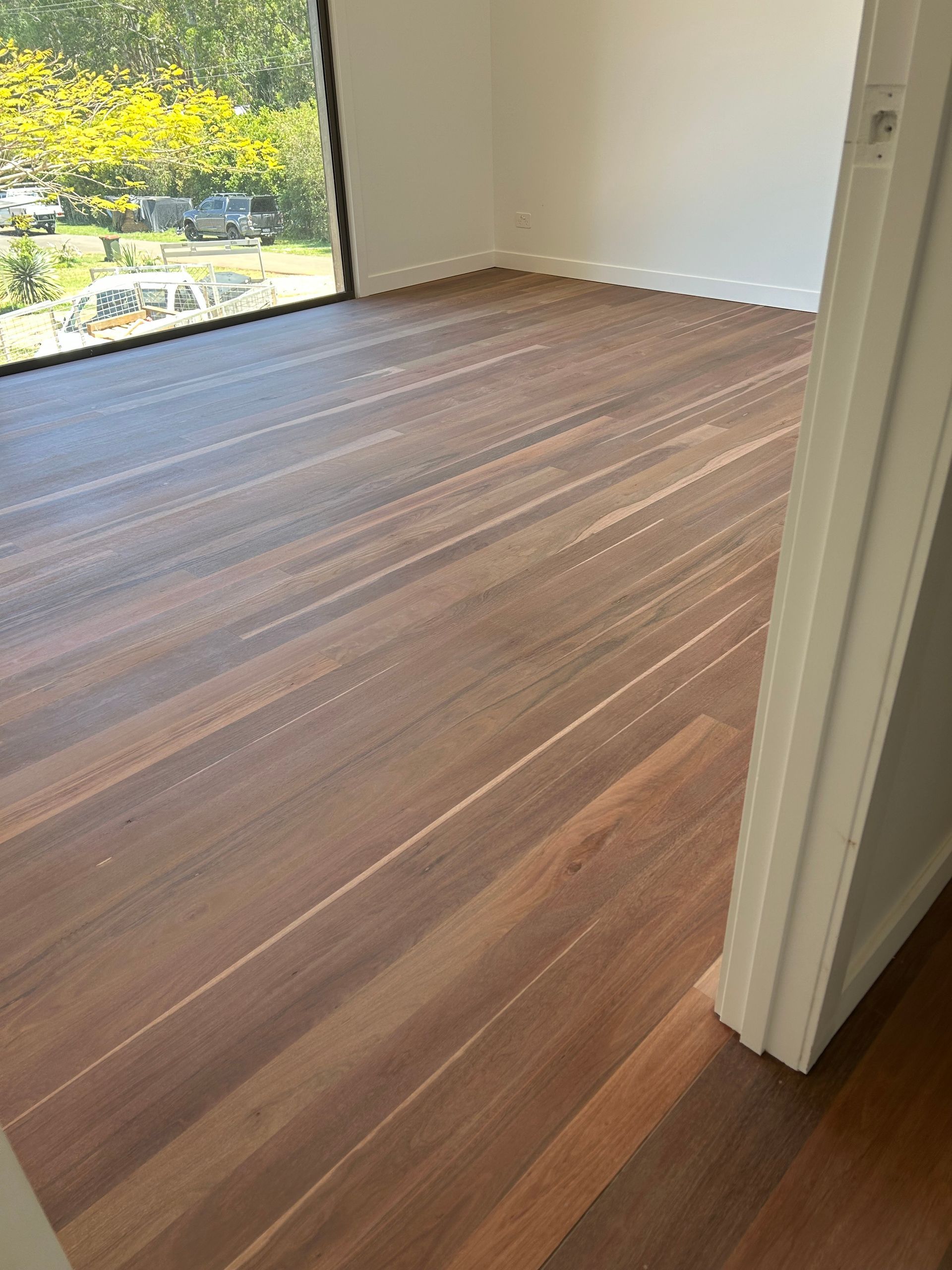 Wooden floor in a room with a large window. The floorboards are a warm brown colour — CM Timber Flooring in East Ballina, NSW