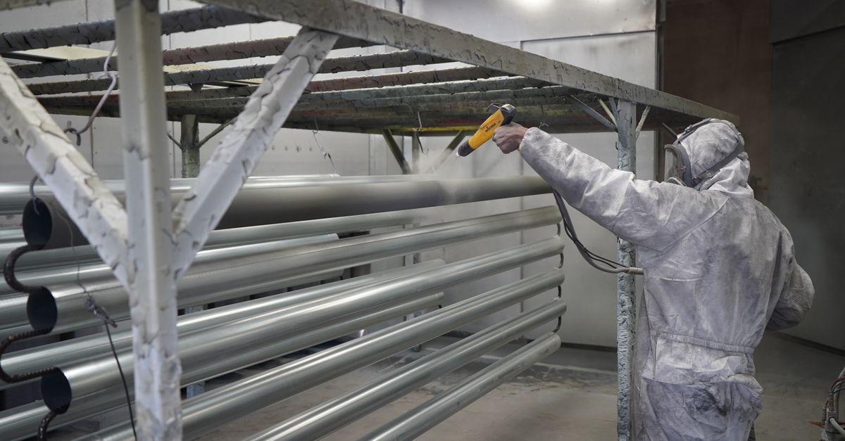 Worker in white protective suit spray-paints metal beams in an industrial facility.