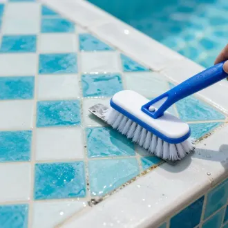 Blue-handled scrub brush cleaning wet blue-and-white pool tiles by the water’s edge