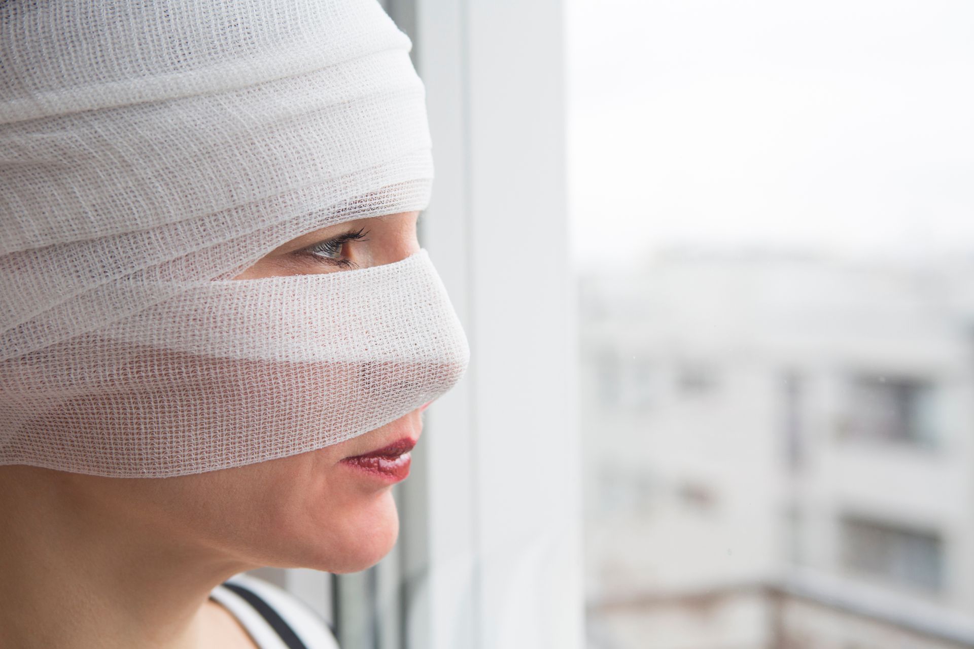 Person with head bandages looking out a window with a blurred building outside