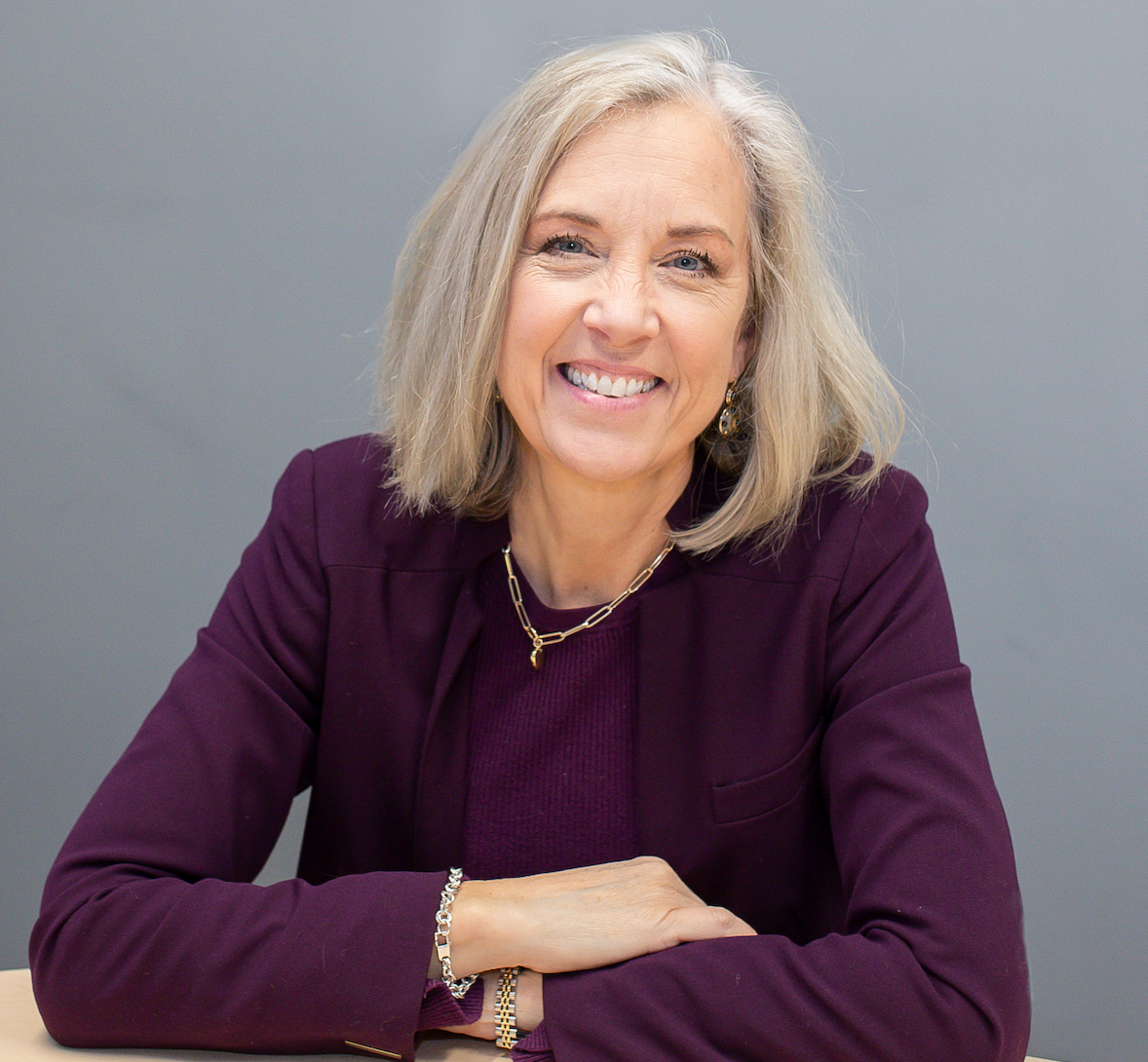 Smiling person in a purple top seated against a gray background, with arms crossed.