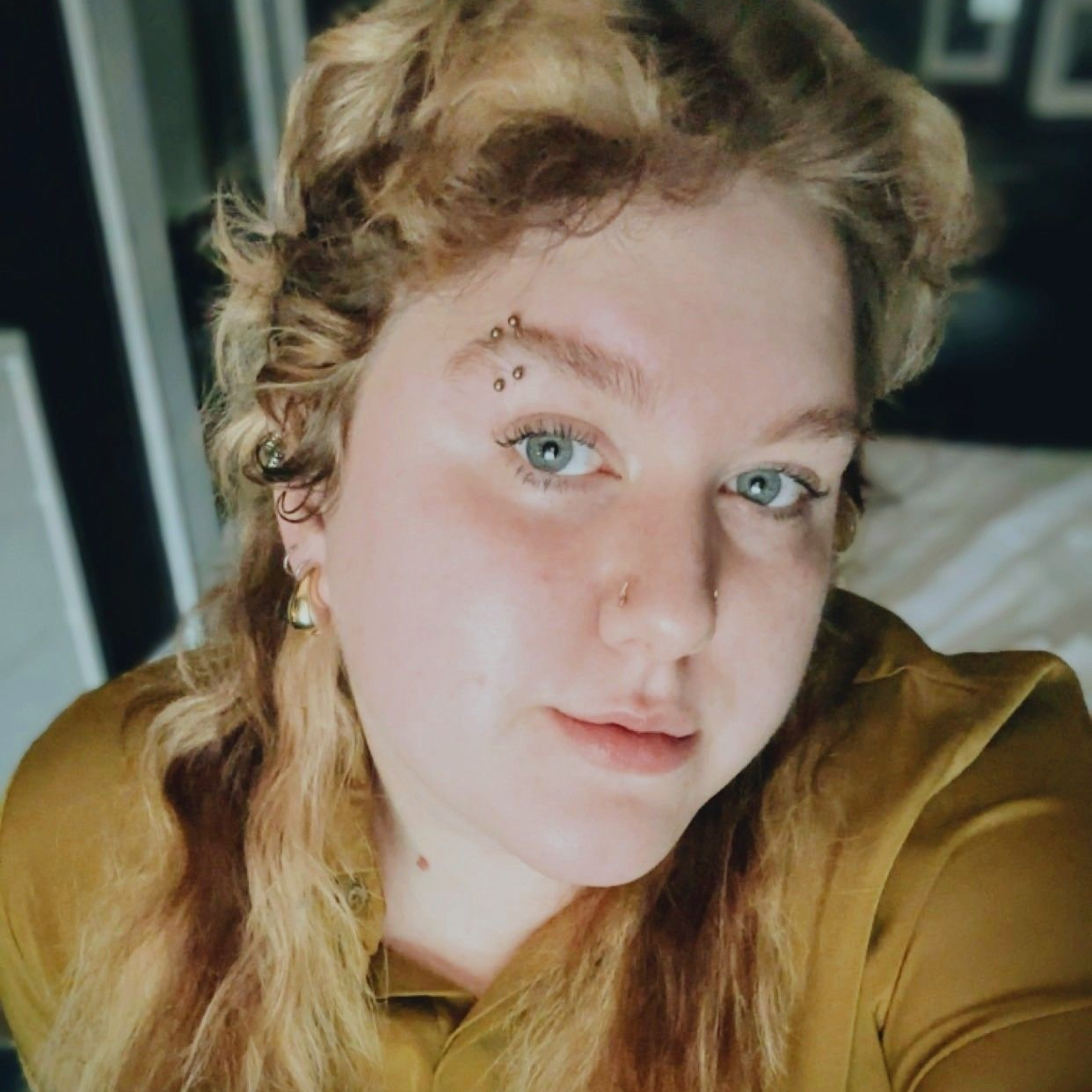 Close-up selfie of a person with wavy blonde hair, blue eyes, and a gold top, indoors
