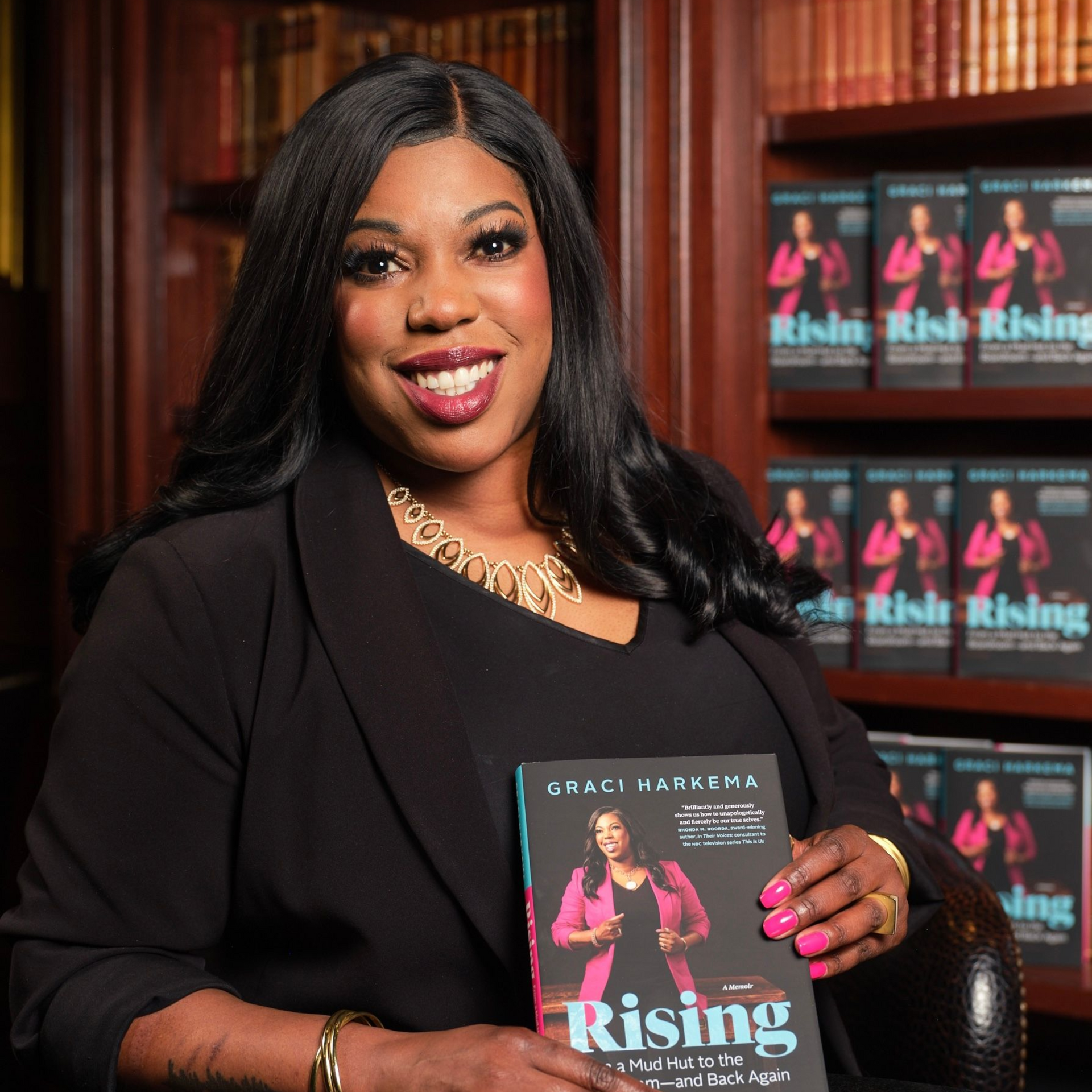 person in black dress holding a book titled “Rising” and smiling in front of matching promotional posters