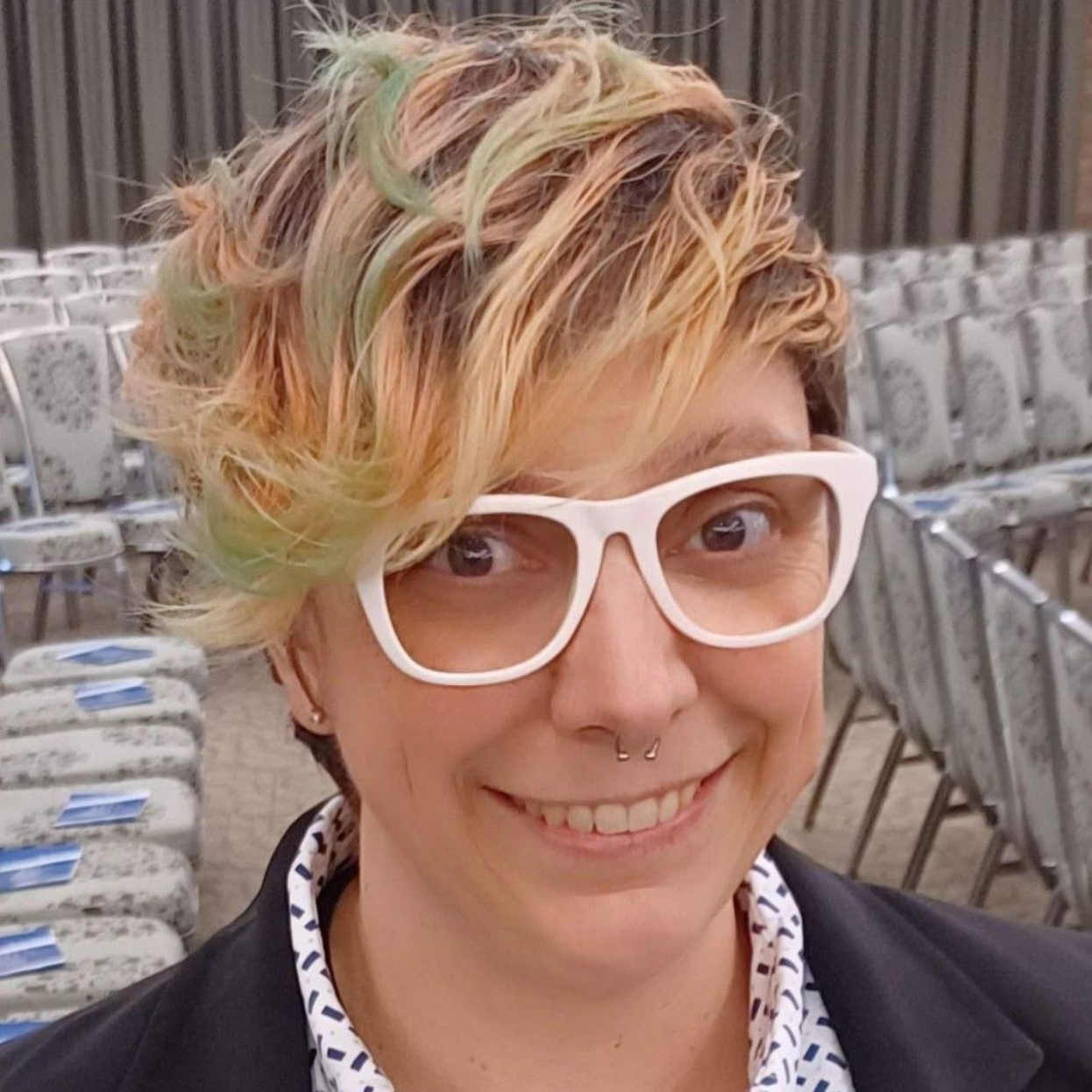 Smiling person with short multicolored hair and white glasses in a room with rows of chairs
