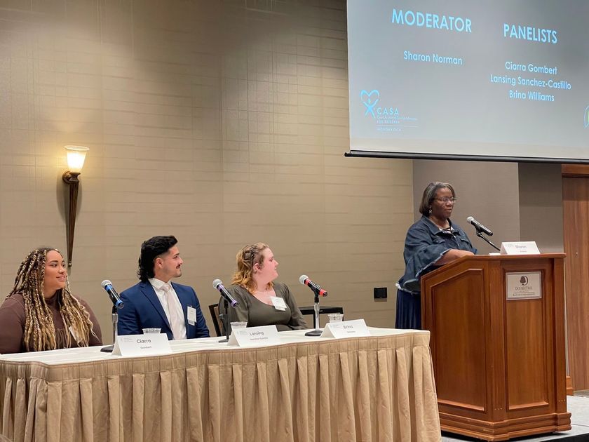 Panel discussion at a conference, with speakers seated at a table and one presenter at a podium on stage.