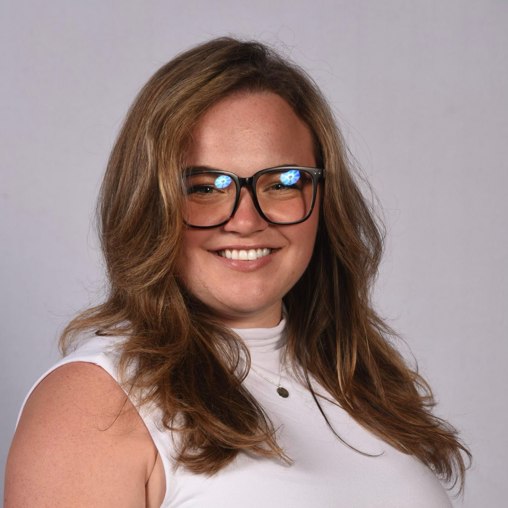 Smiling person with long brown hair and glasses, wearing a white top against a plain background