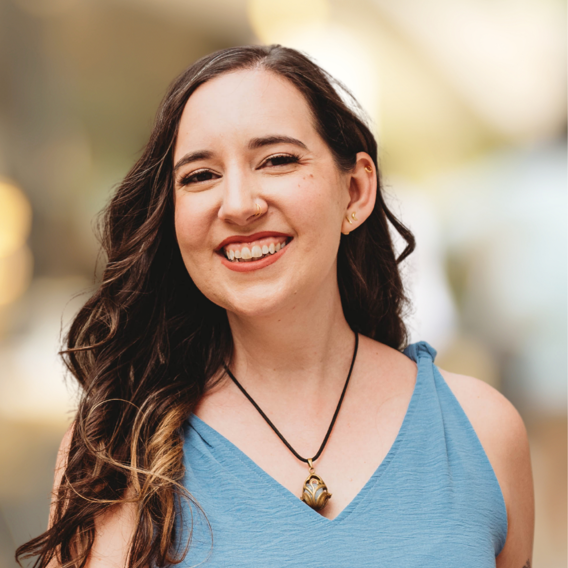 Smiling person with long dark hair wearing a blue top and pendant necklace outdoors with blurred background