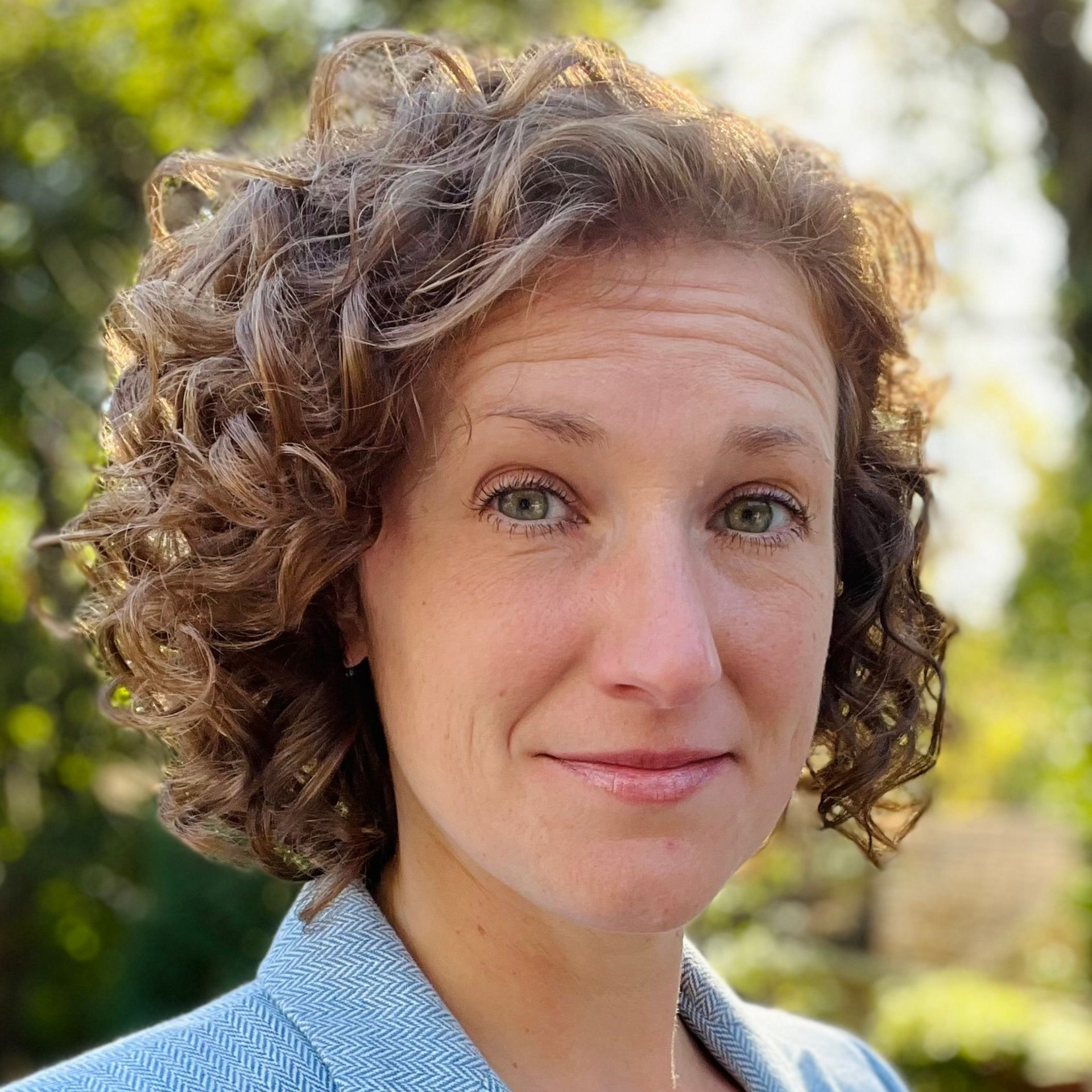 Close-up portrait of a smiling person with curly hair in a sunlit outdoor garden