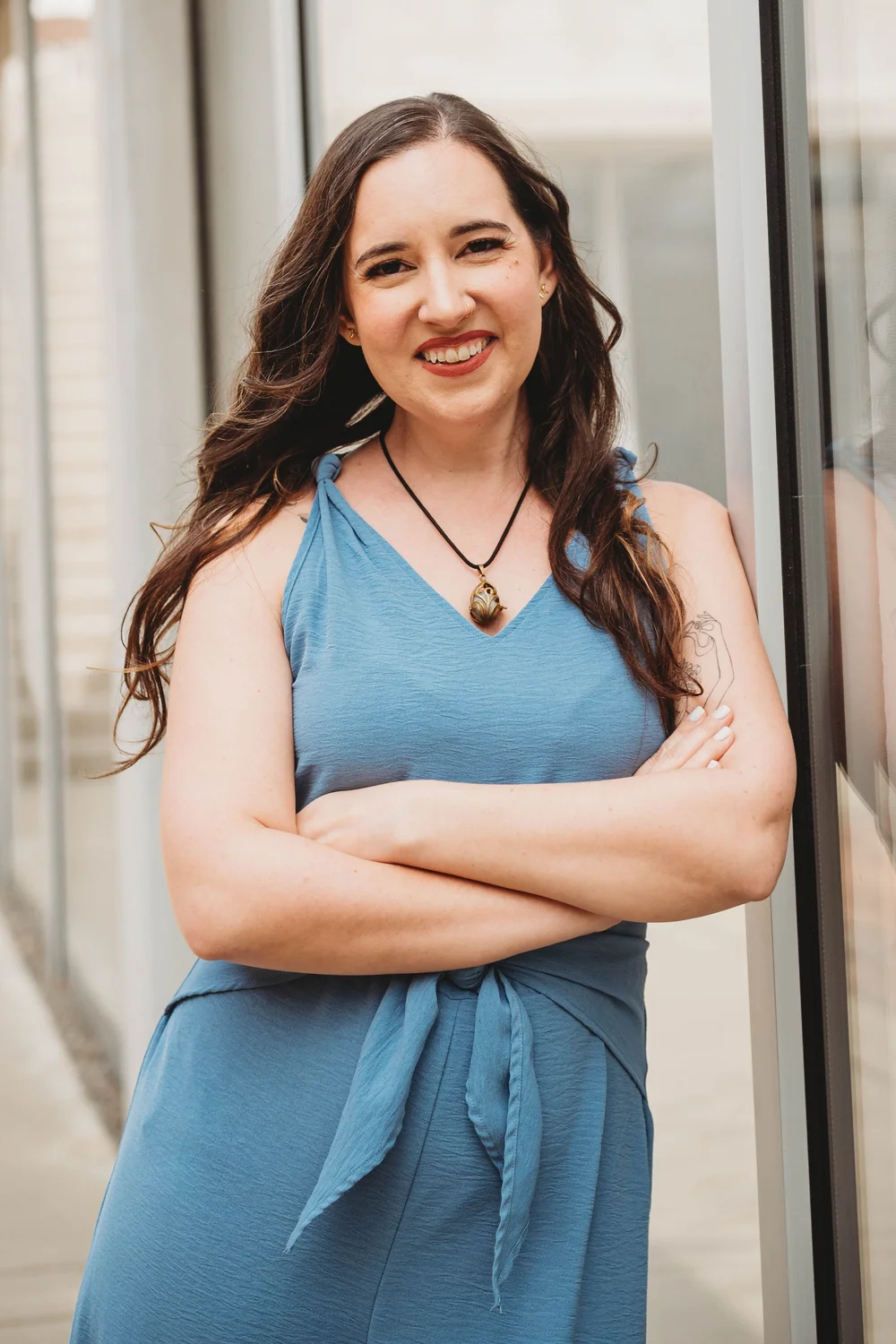 Smiling person in a blue dress with arms crossed, standing outdoors by a window
