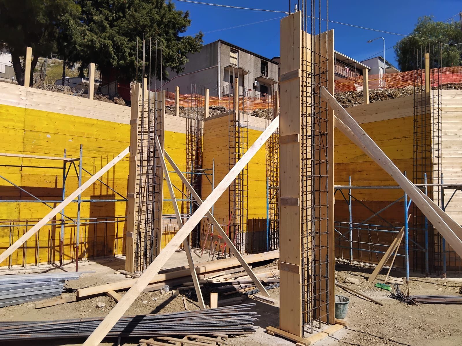 Cantiere con casseforme in legno per pilastri in calcestruzzo e muro di contenimento, sostenuti da supporti.
