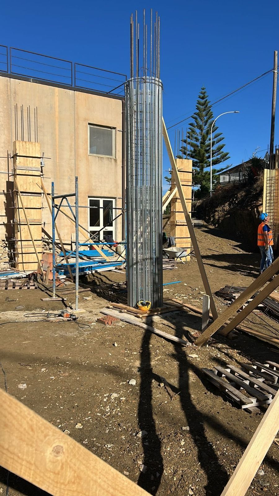 Cantiere con un'alta colonna di metallo e barre di armatura, edifici circostanti e operai edili.