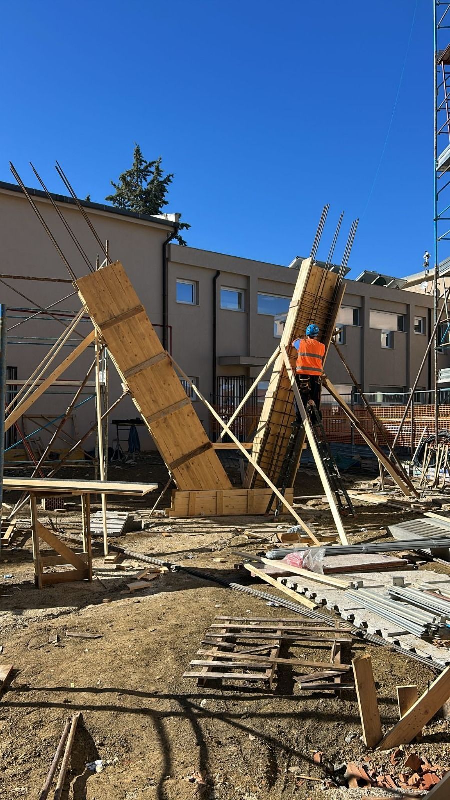 Cantiere con due forme di legno angolate, sostenute da rinforzi; un operaio con il gilet arancione.
