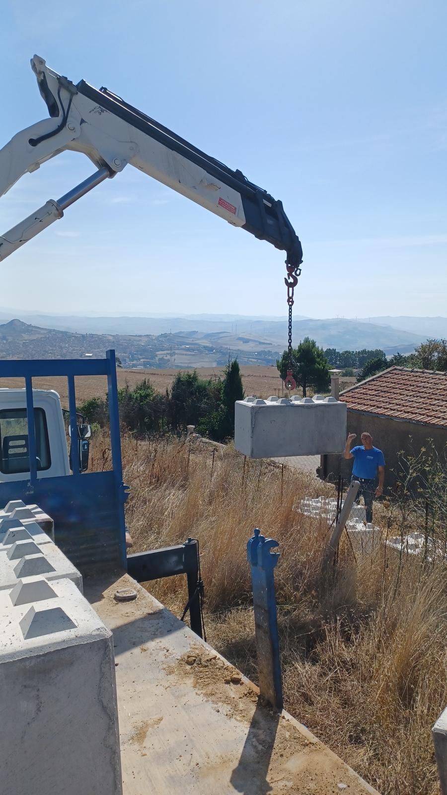 Una gru solleva un blocco di cemento dal cassone di un camion. Un uomo osserva vicino a una casa in una giornata di sole.