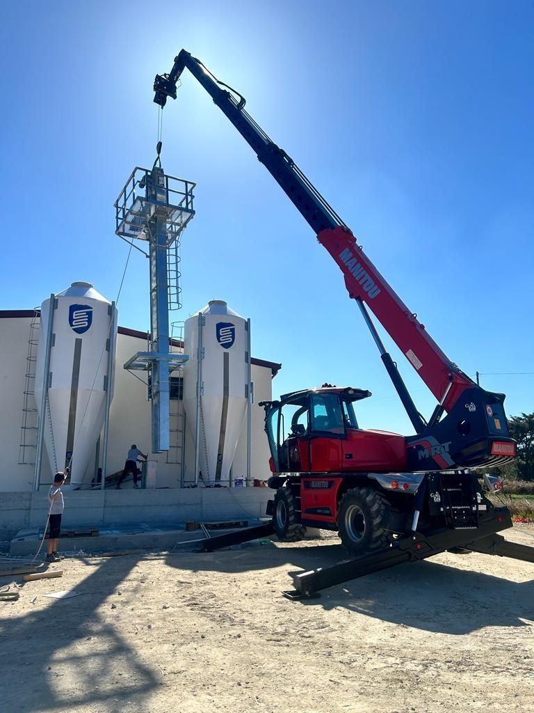 Sollevatore telescopico rosso che solleva una struttura metallica vicino ai silos per cereali in un cantiere edile sotto un cielo soleggiato.