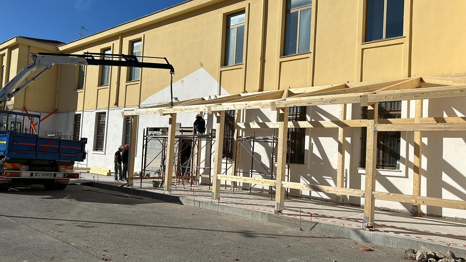 Cantiere: operai che costruiscono un pergolato in legno attaccato a un edificio giallo. Presente un camion con gru.