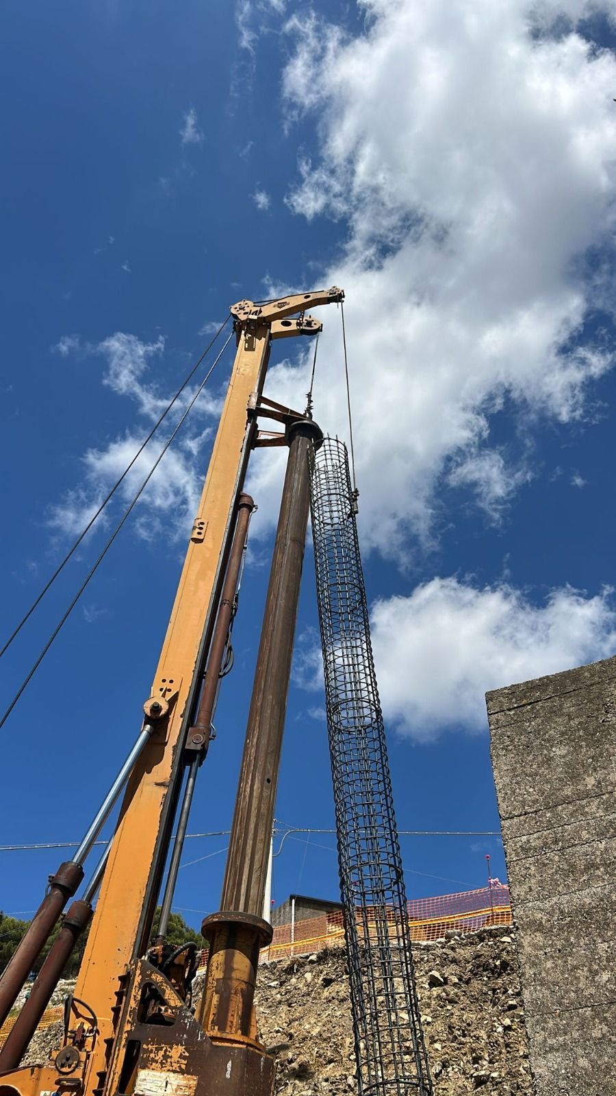 Gru da costruzione gialla che perfora il terreno, cielo azzurro e muro di pietra.