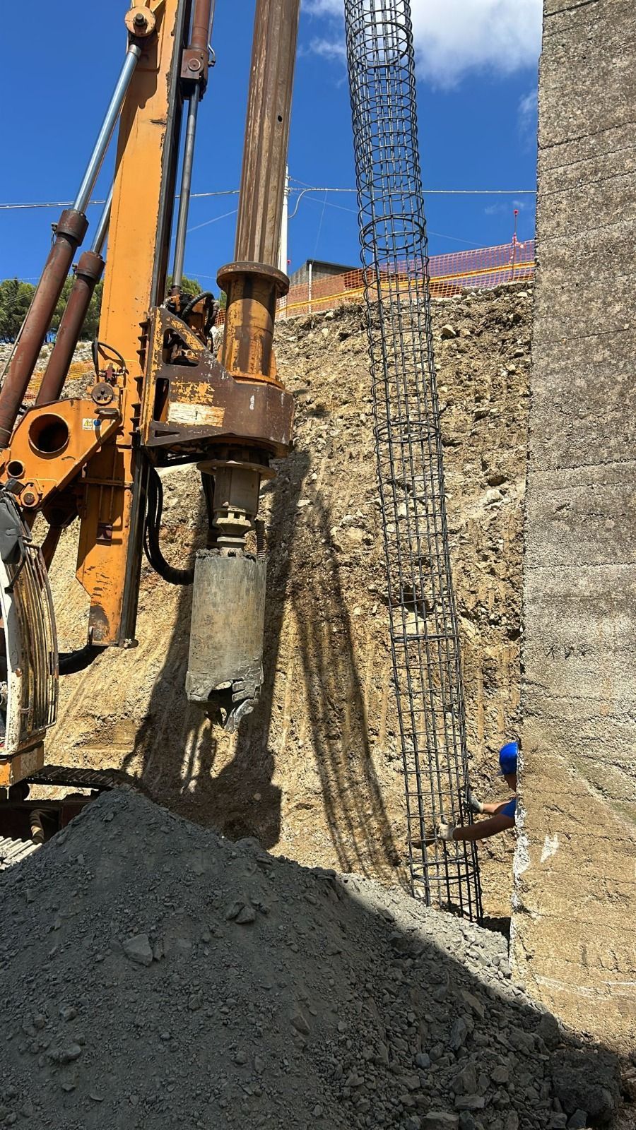 Cantiere edile: perforatrice che perfora il terreno, cumulo di terra in primo piano, barre di armatura e operai sullo sfondo.