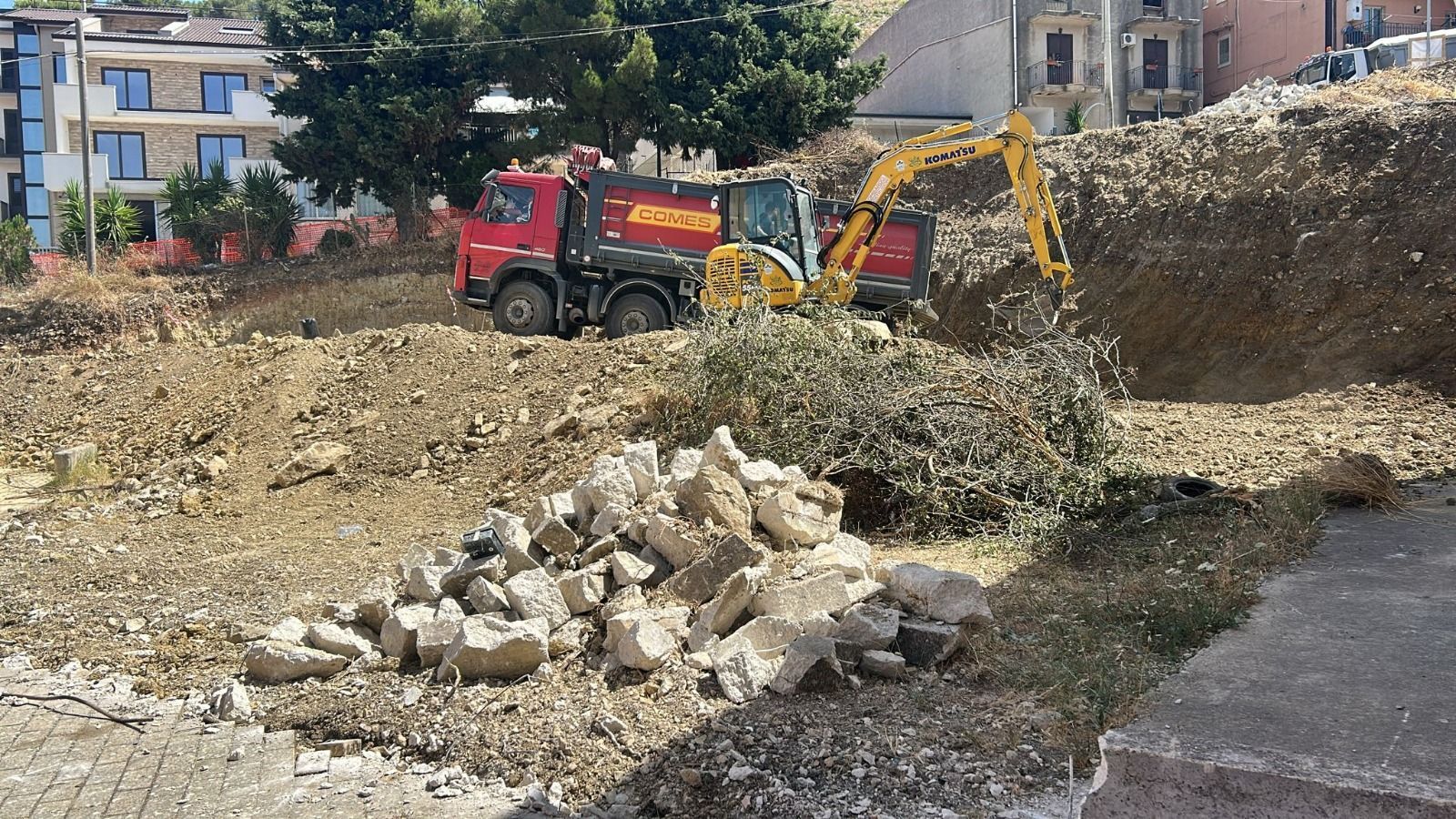 Un escavatore carica terra su un autocarro con cassone ribaltabile rosso in un cantiere edile vicino agli edifici.