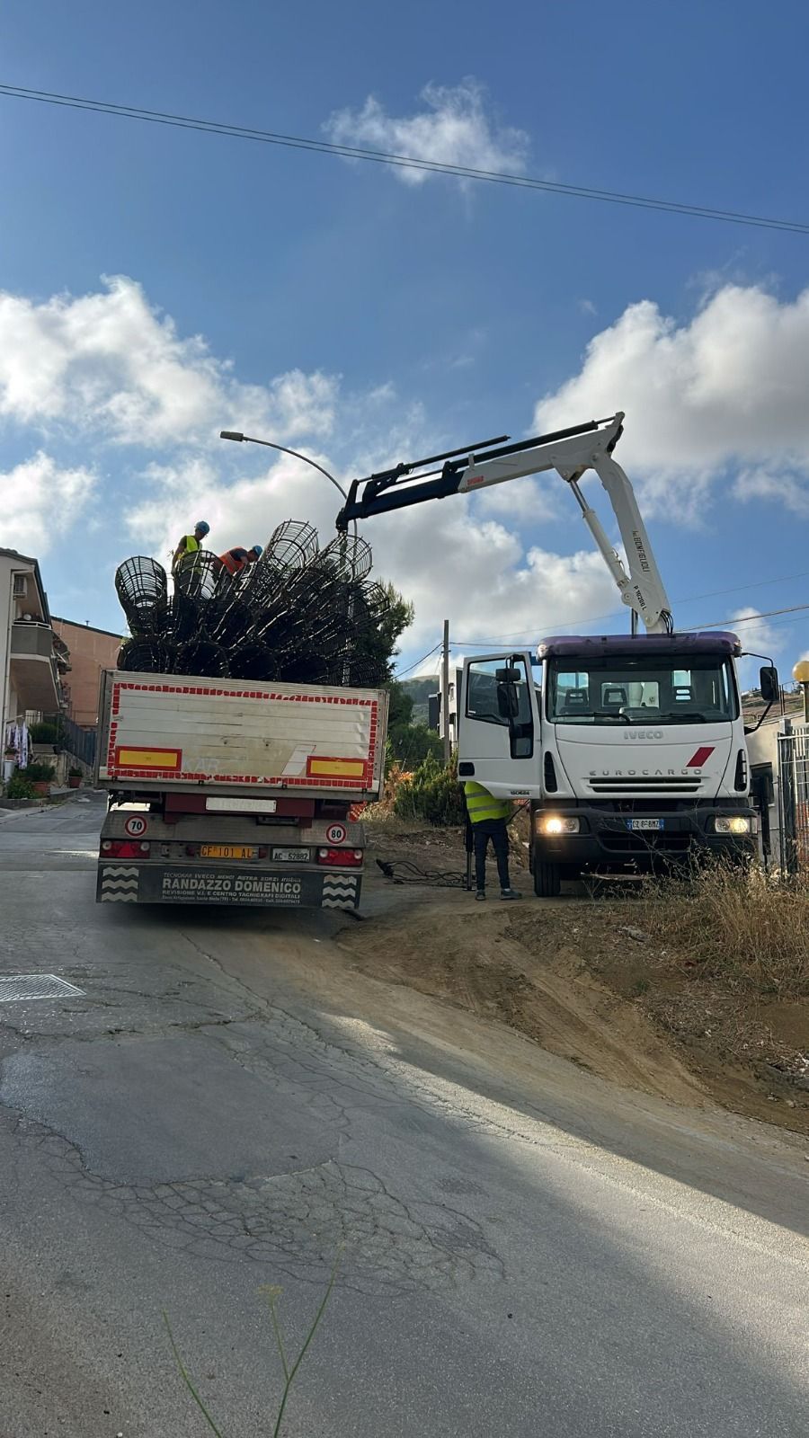 Un camion carica detriti con una gru su una strada dissestata, con edifici sullo sfondo e cielo azzurro.