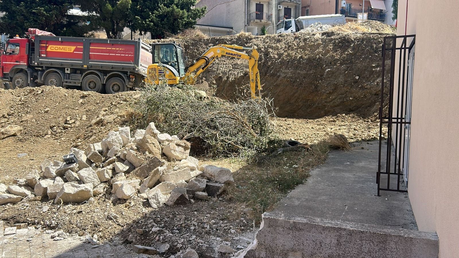 Un escavatore carica terra e detriti su un autocarro con cassone ribaltabile in un cantiere edile.