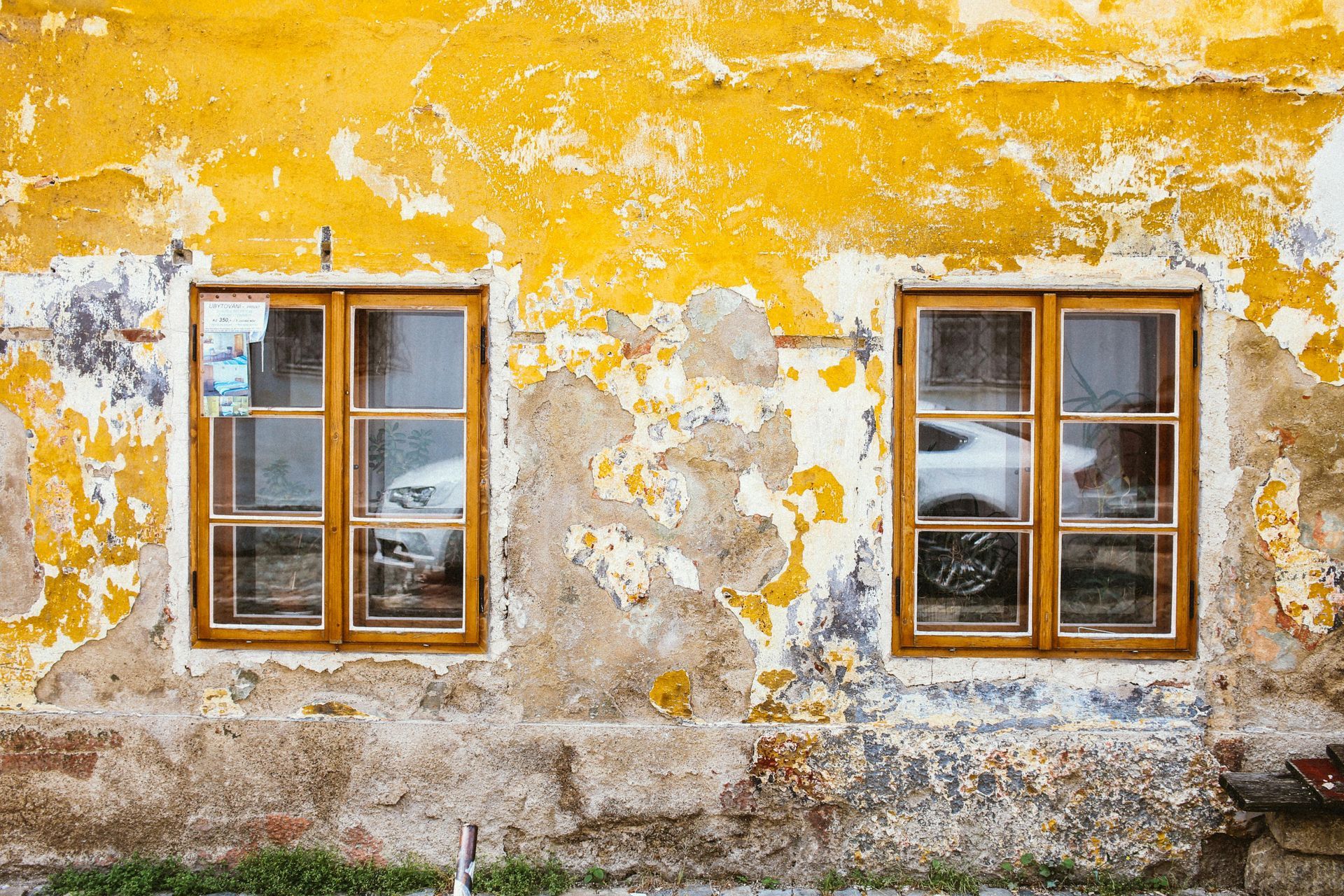 A yellow cracked paint on a wall with two windows and a car parked in front of it.
