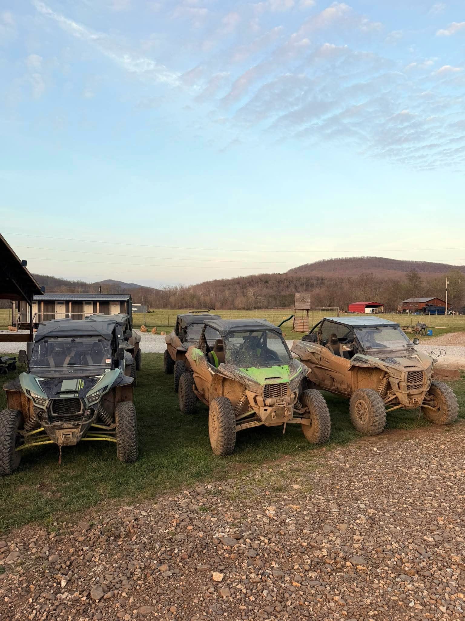 Three muddy off-road vehicles parked on a gravel lot at dusk with hills in the background.