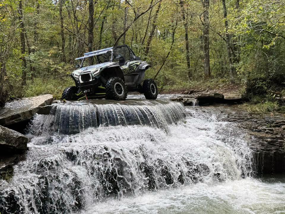 Off-road buggy perched on a rocky waterfall in a wooded forest.