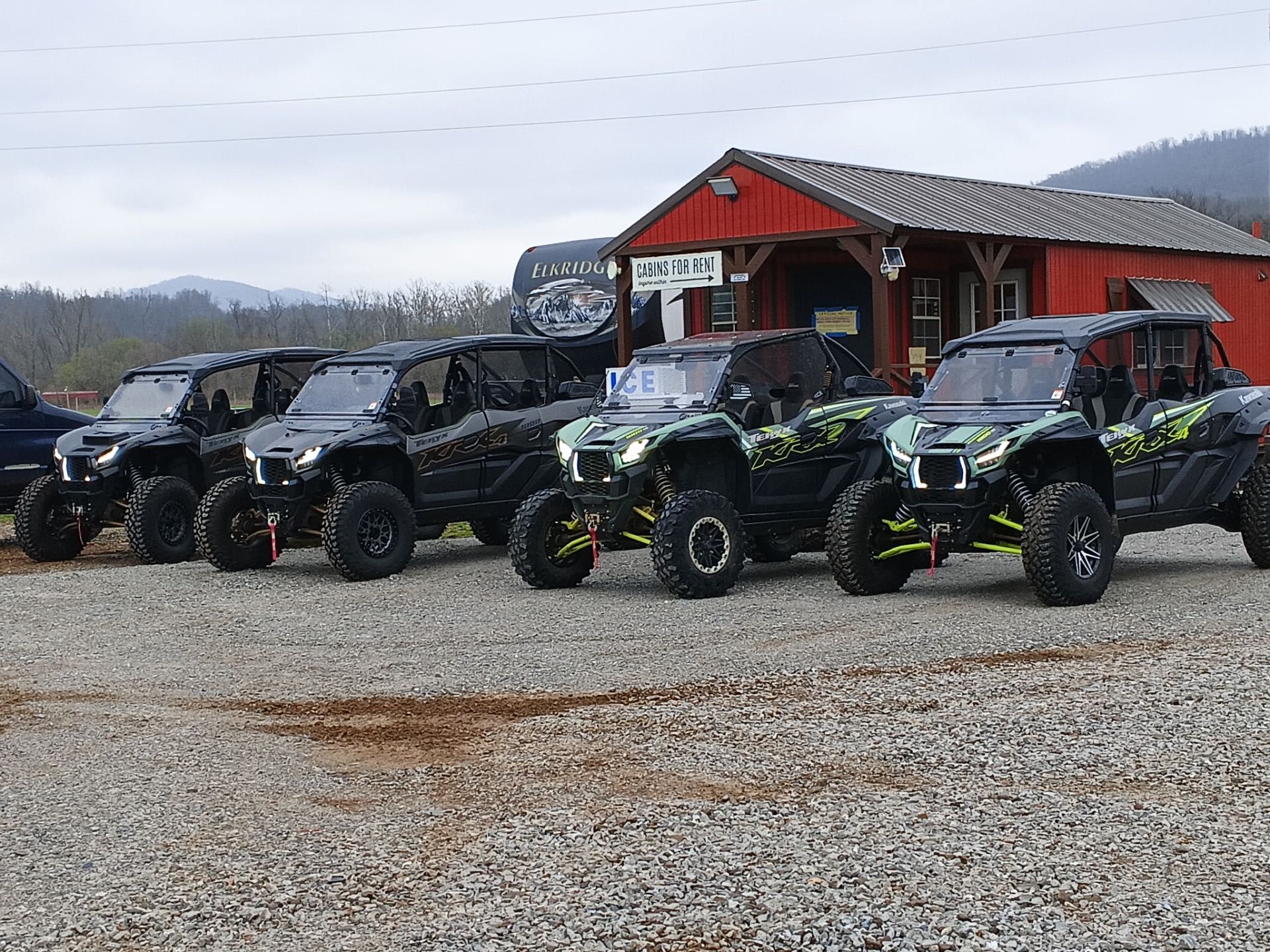 Off-road side-by-side vehicles lined up in front of a red building on a gravel lot, with mountains in the background