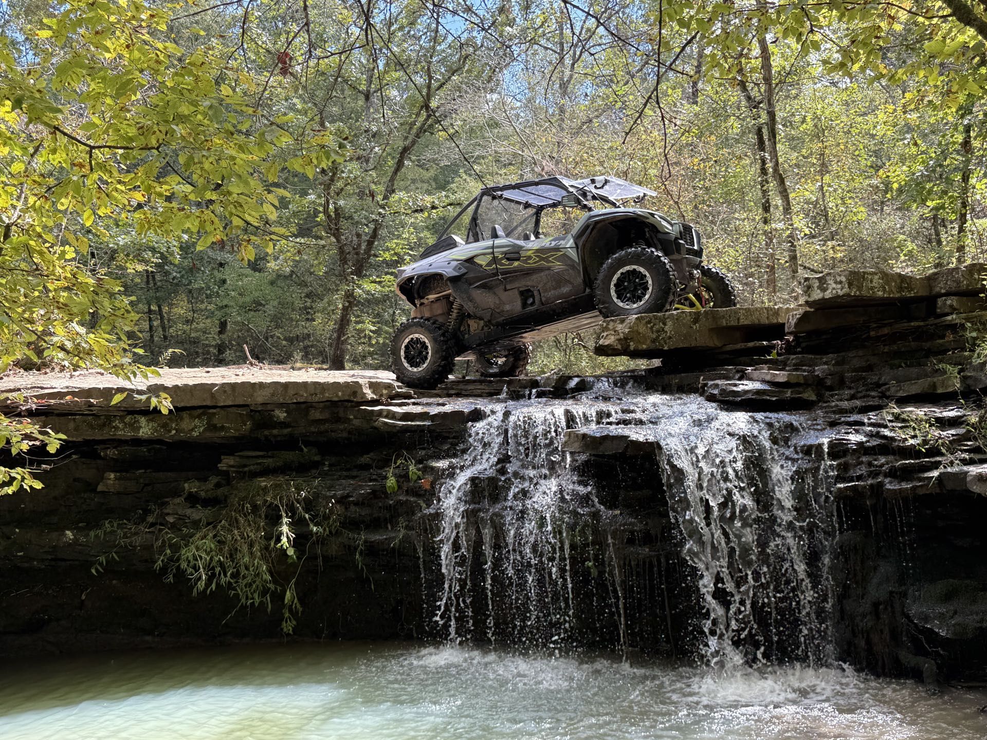 Off-road buggy crossing a rocky ledge above a small waterfall in a wooded area
