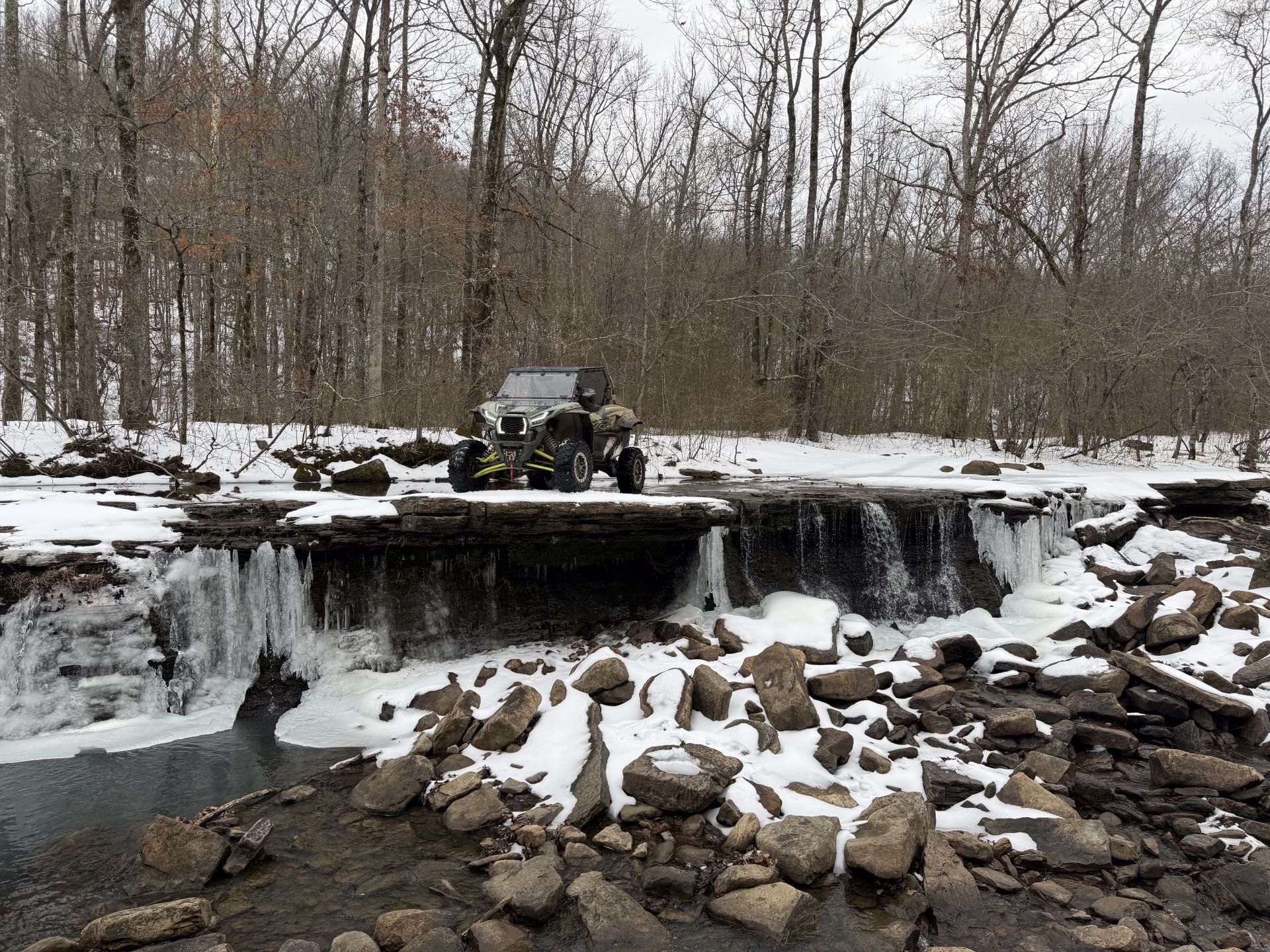 Snowy waterfall in a wooded area with rocks and a small vehicle on the cliff above
