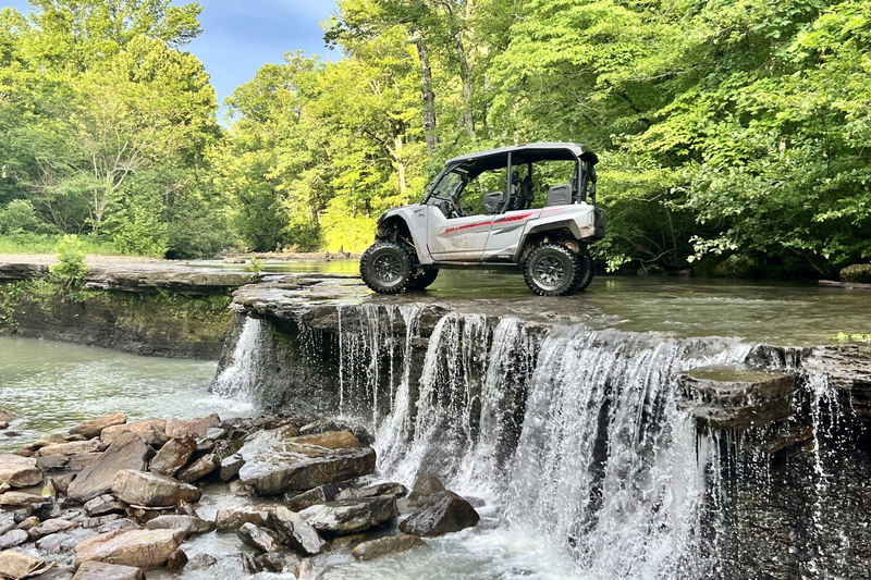 Off-road vehicle perched on a small waterfall in a wooded creek.