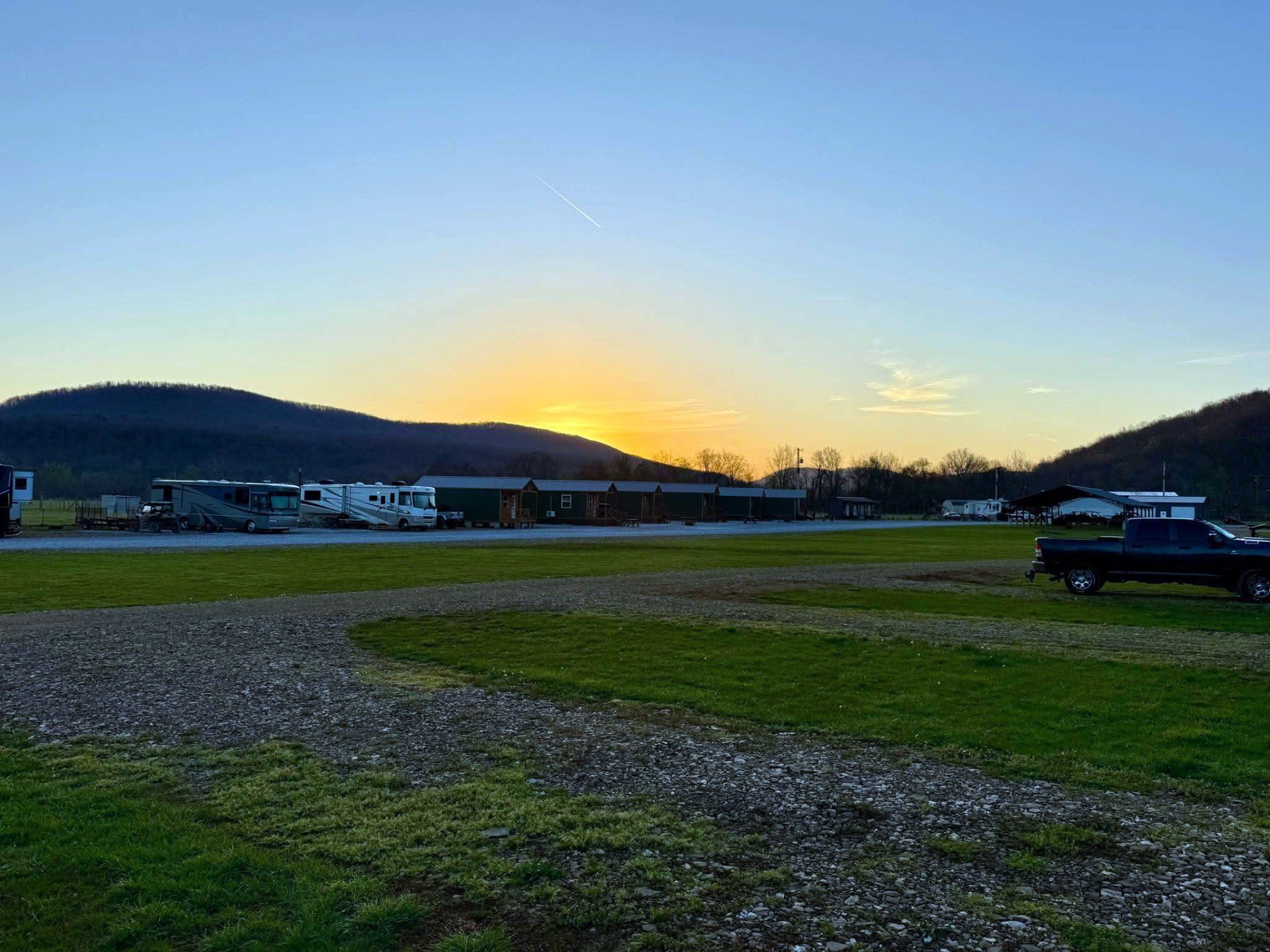 Sunset over a grassy field with hills, parked cars, and RVs under a clear blue sky