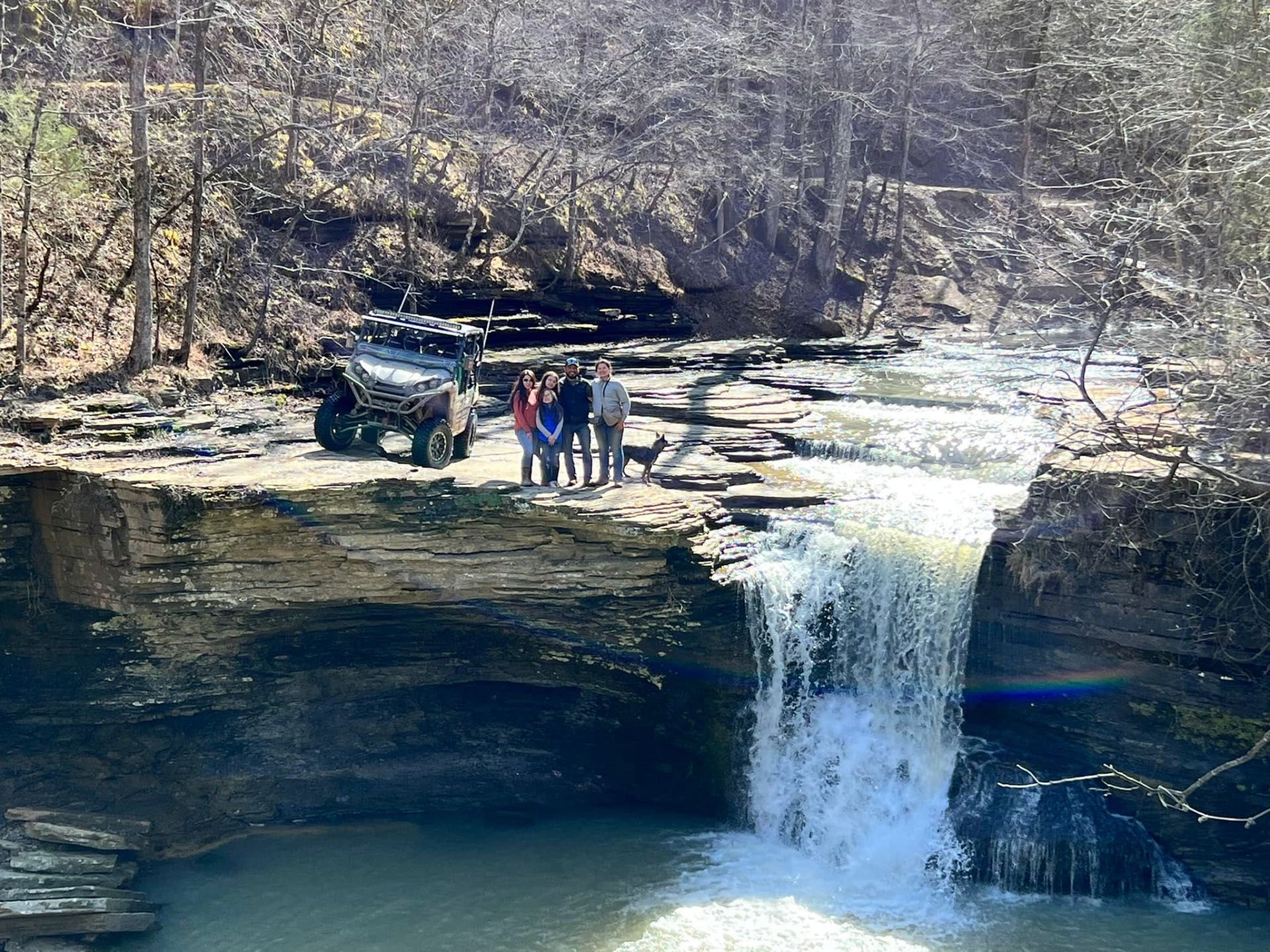 People standing by a waterfall beside an off-road vehicle in a wooded ravine