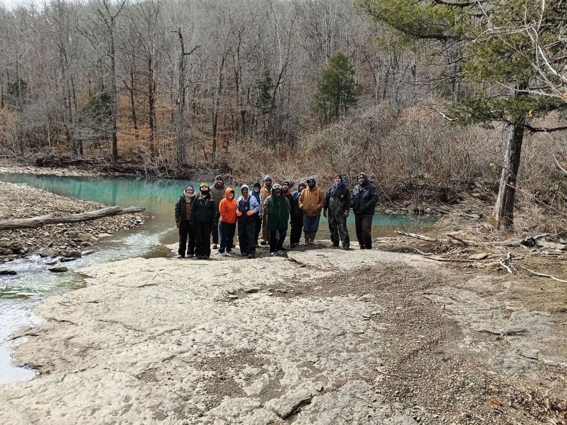 Group of people standing on rocky ground beside a turquoise river in a leafless forest