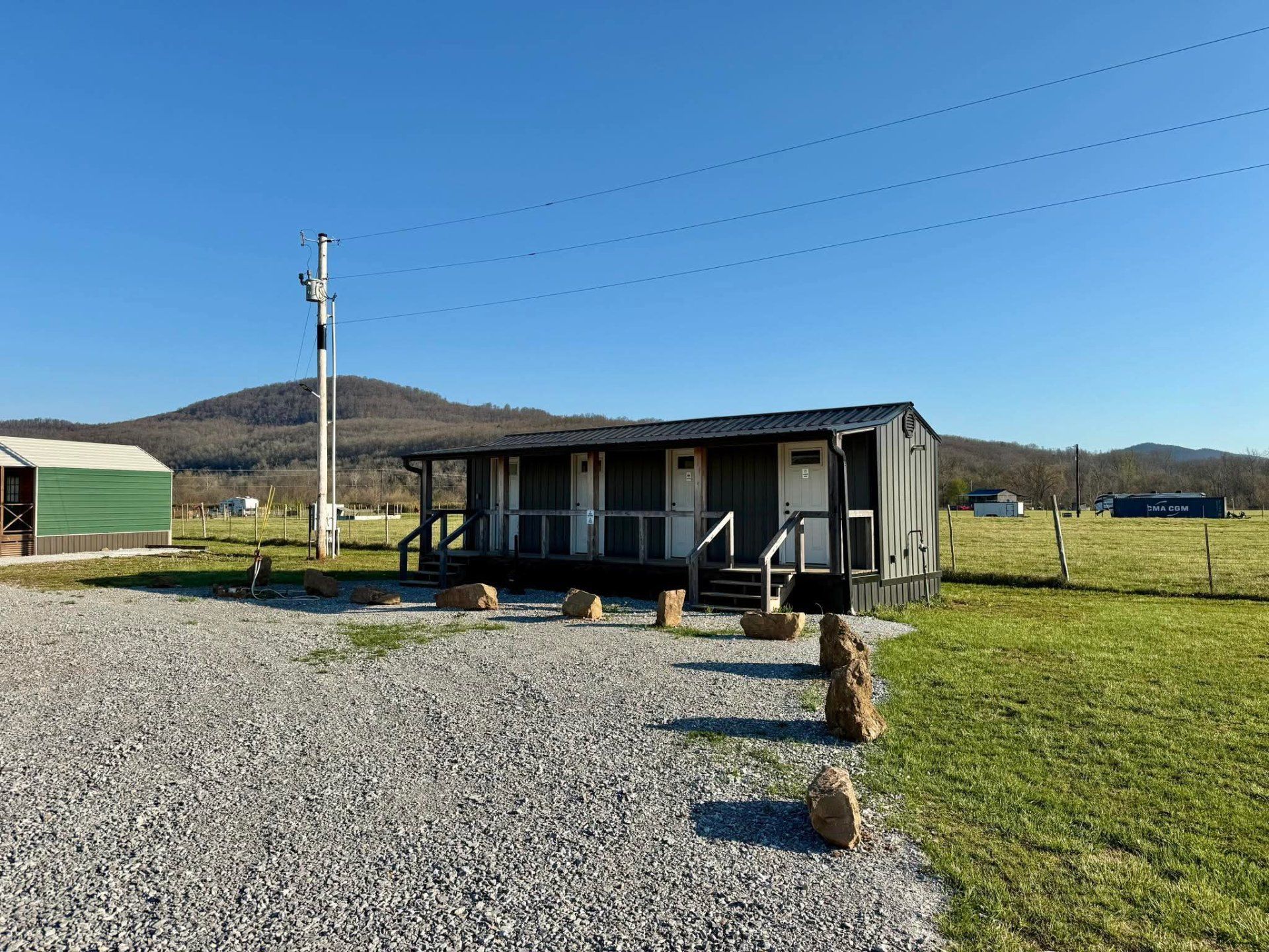 Small rustic cabin on gravel in a grassy rural field with hills and blue sky.