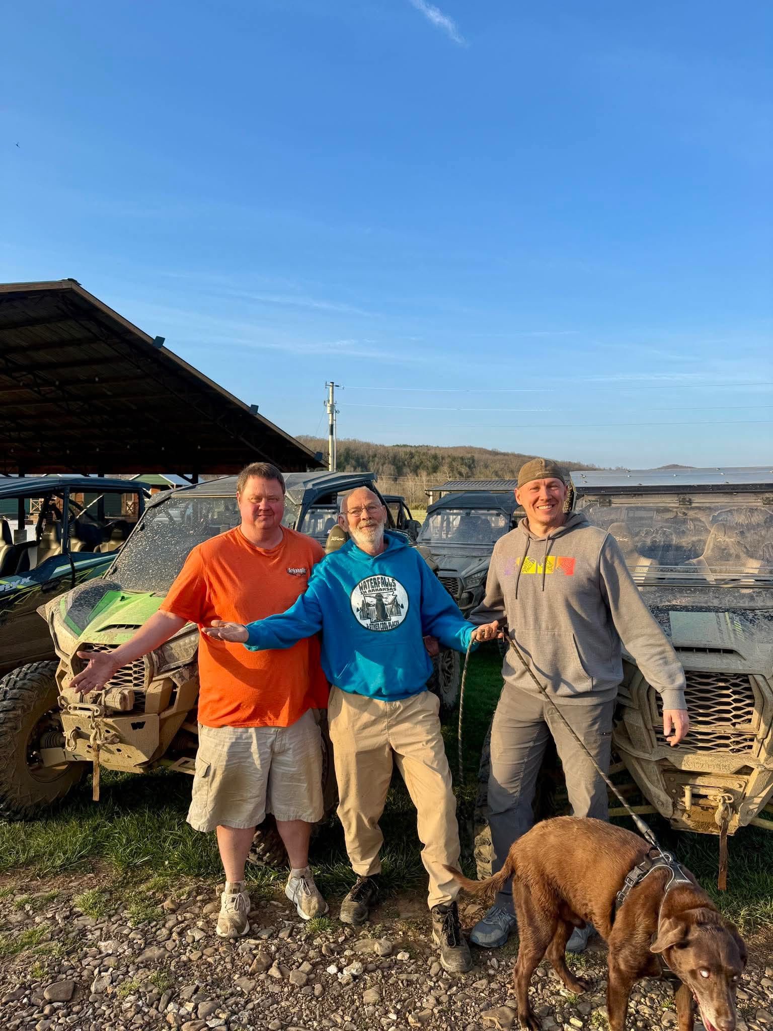 Three people stand with a dog beside a muddy off-road vehicle under a shelter near water.