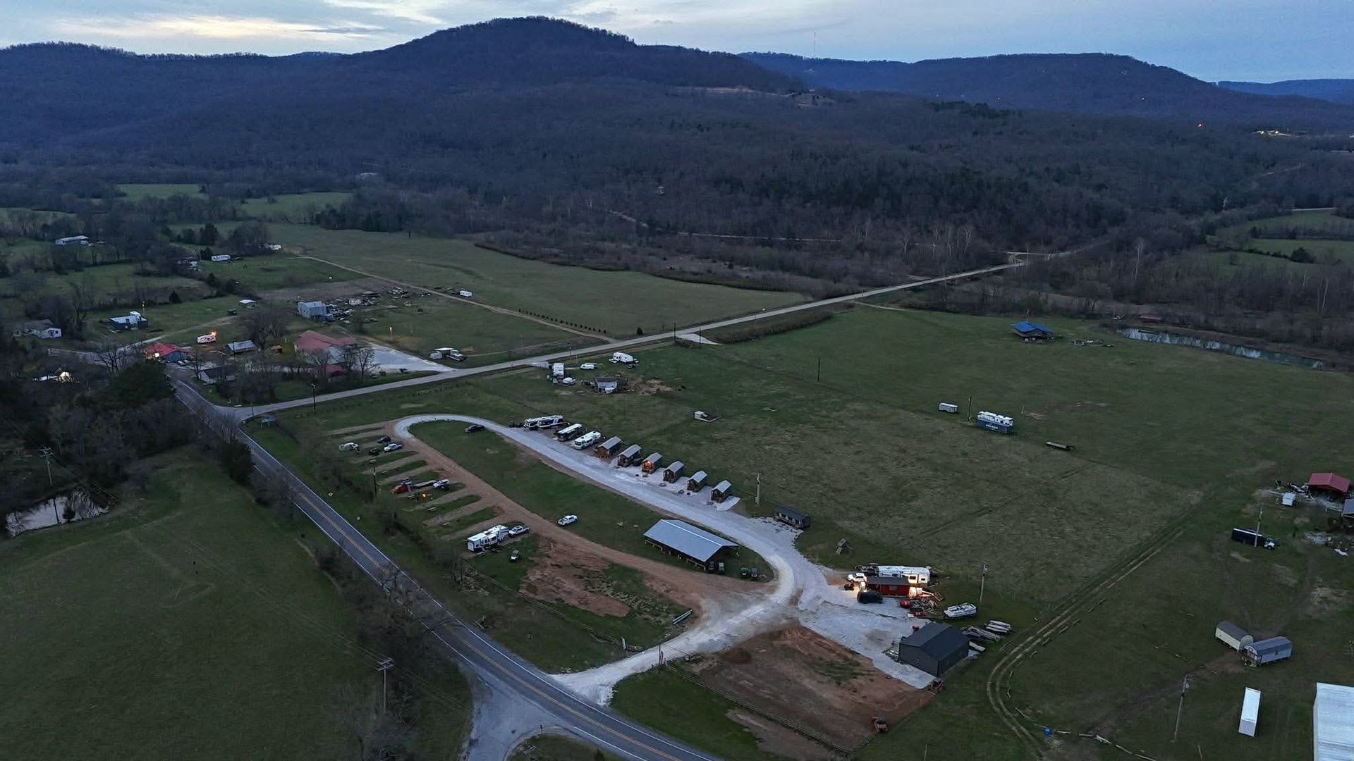 Aerial view of a rural campground with RVs, roads, and fields beside wooded hills at dusk