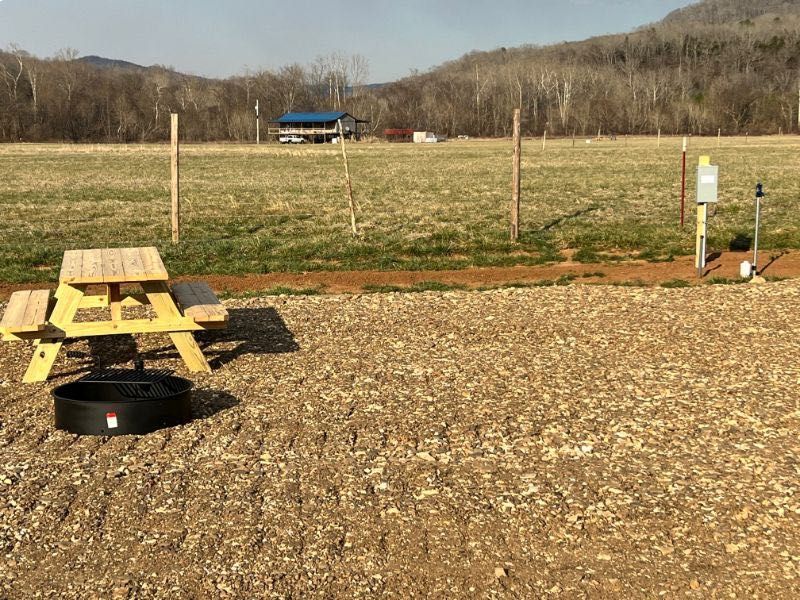 Gravel lot with a yellow picnic table, fire ring, and open field with a barn in the distance