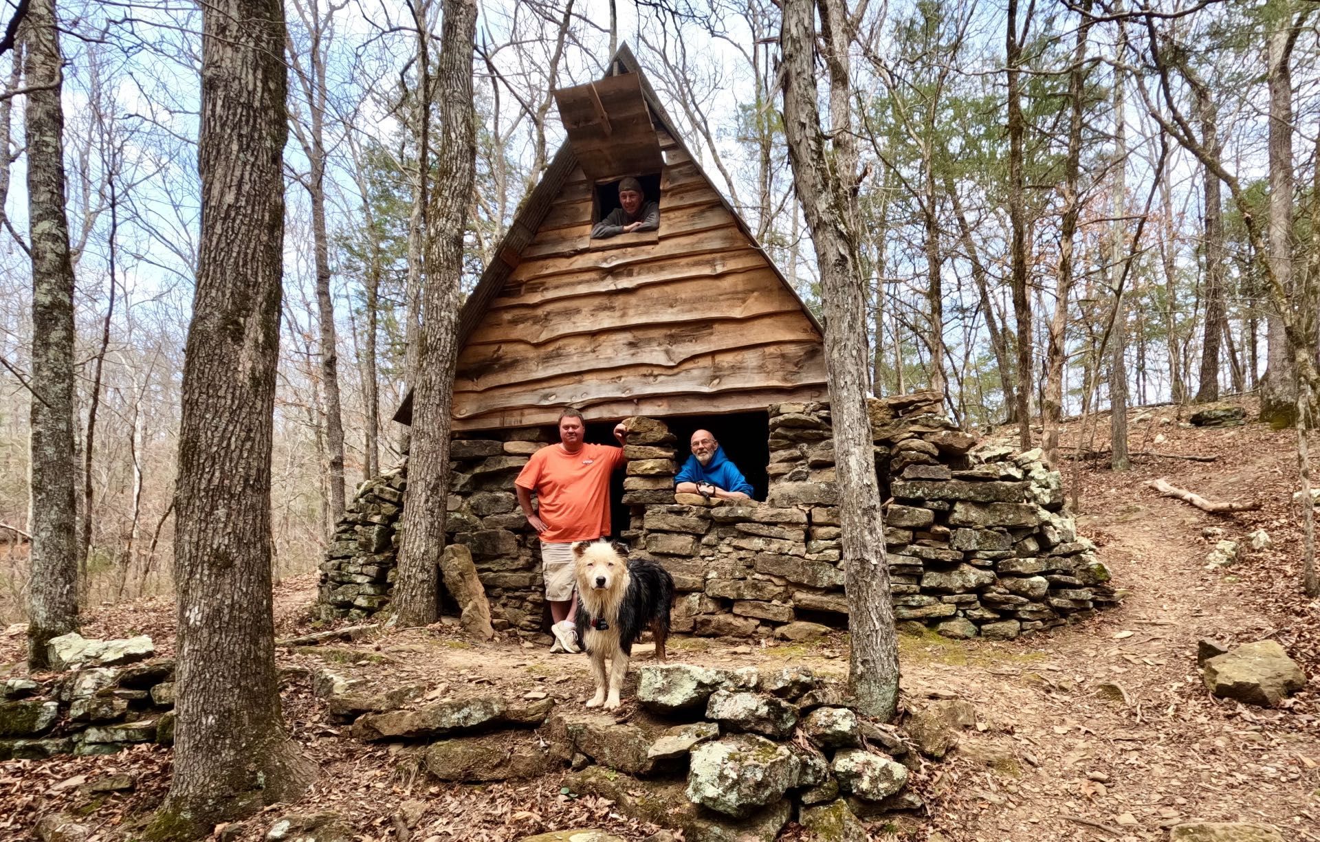 Two hikers with a dog standing by a small wooden shelter in the woods